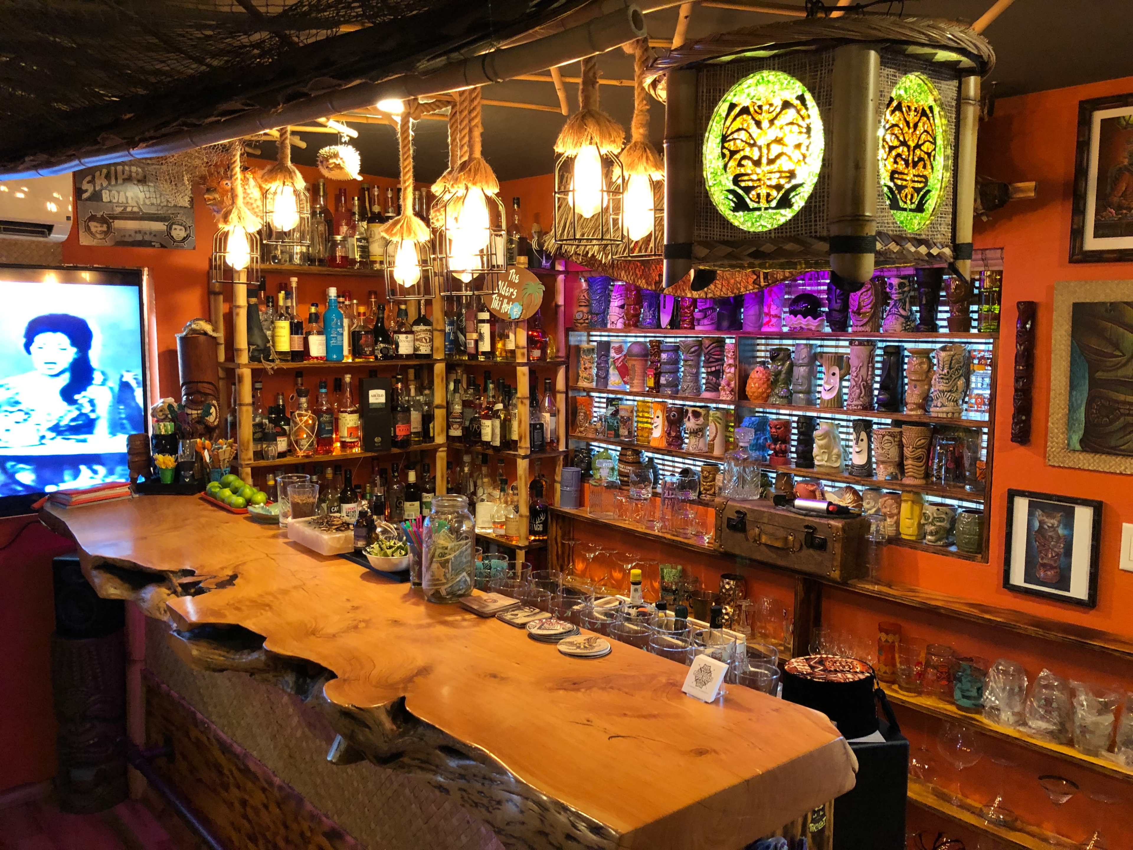 The image shows a vibrant bar with an extensive collection of bottles and tiki mugs arranged behind a wooden counter, illuminated by colorful lighting and decorative tiki-themed elements.