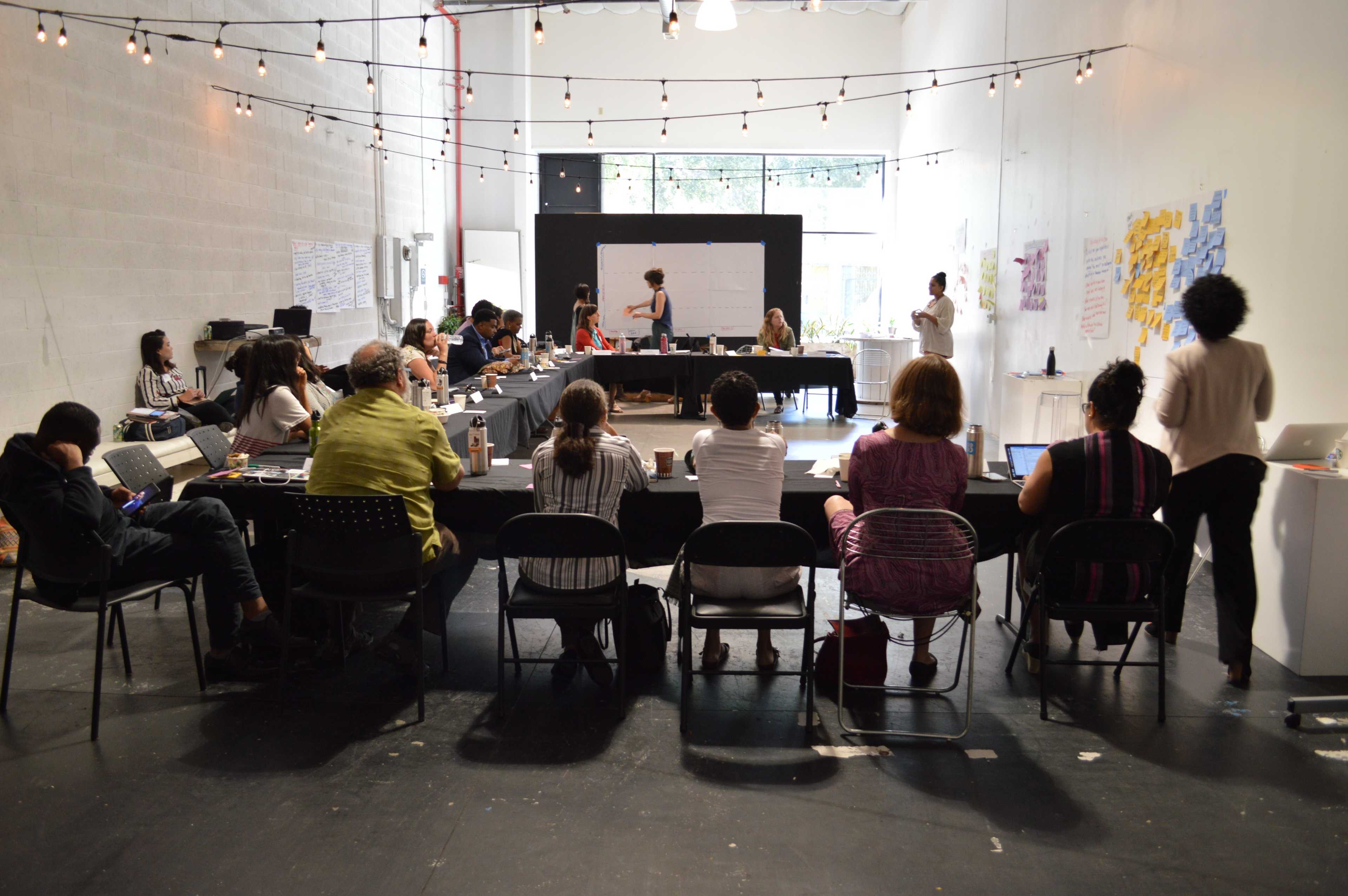 A group of people is seated at a long table in a bright, airy room, engaged in a meeting while a presenter stands in front of a whiteboard.
