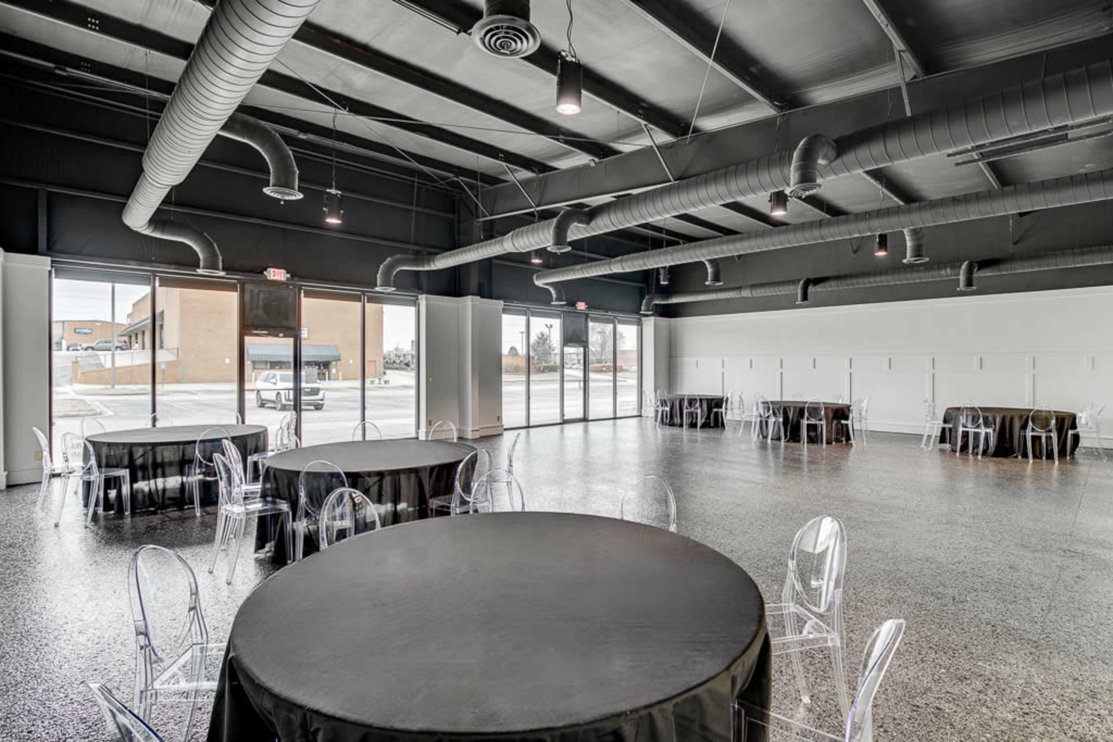 The interior of an event space features several round tables with black tablecloths and transparent chairs, illuminated by natural light from large windows.