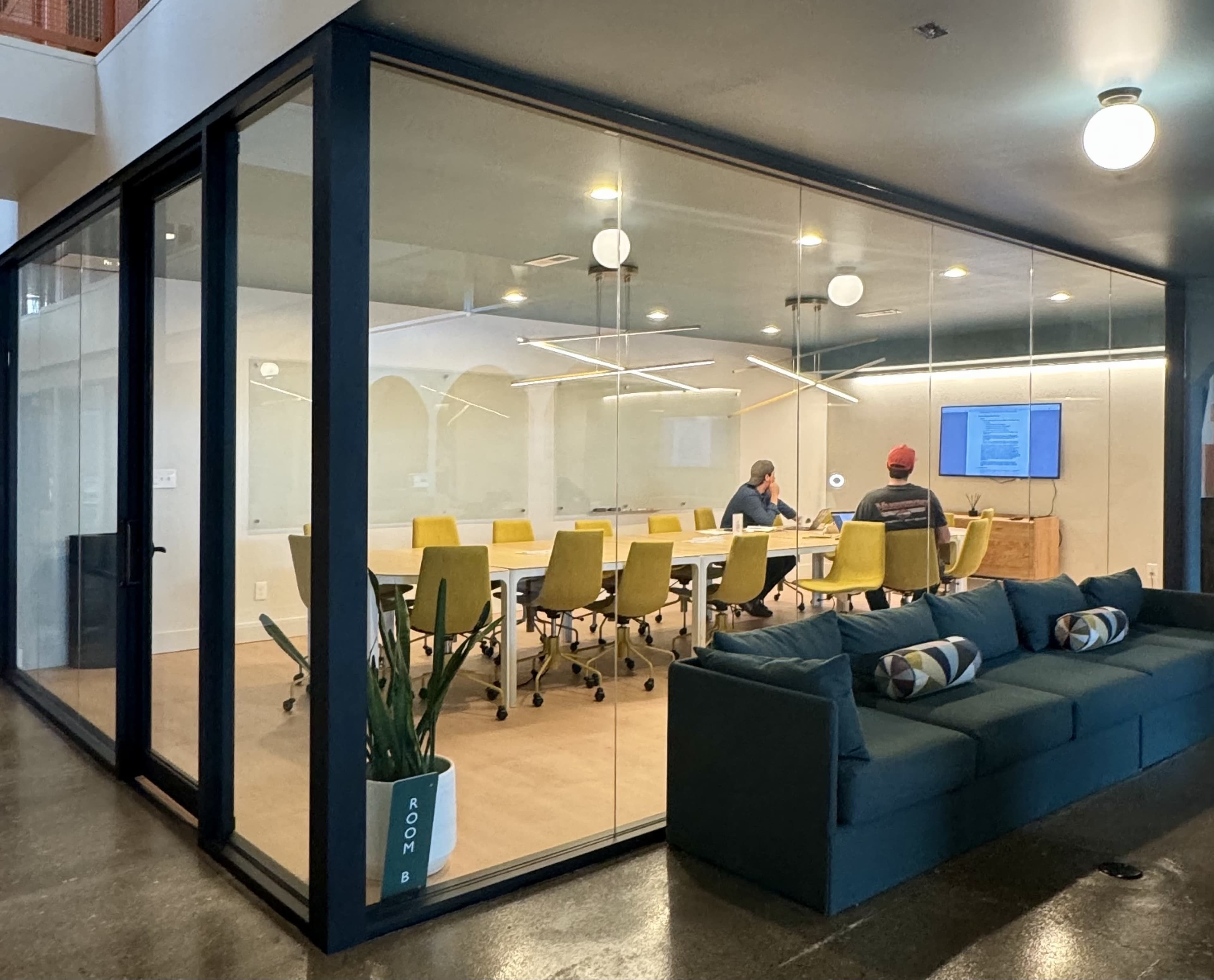 A modern conference room with glass walls features a long table surrounded by yellow chairs, while two people work at a computer in the foreground.