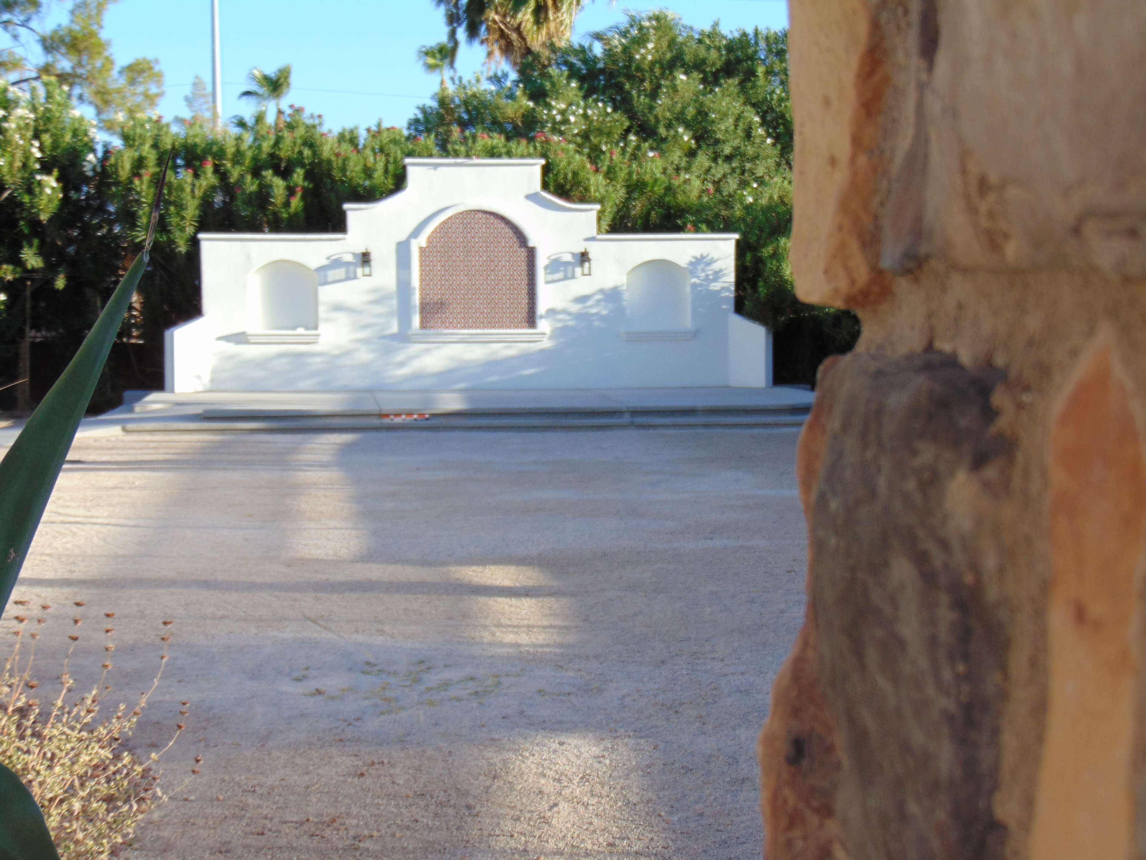 The image features a white, architectural structure with a decorative central panel, flanked by two small alcoves, set against a backdrop of greenery.