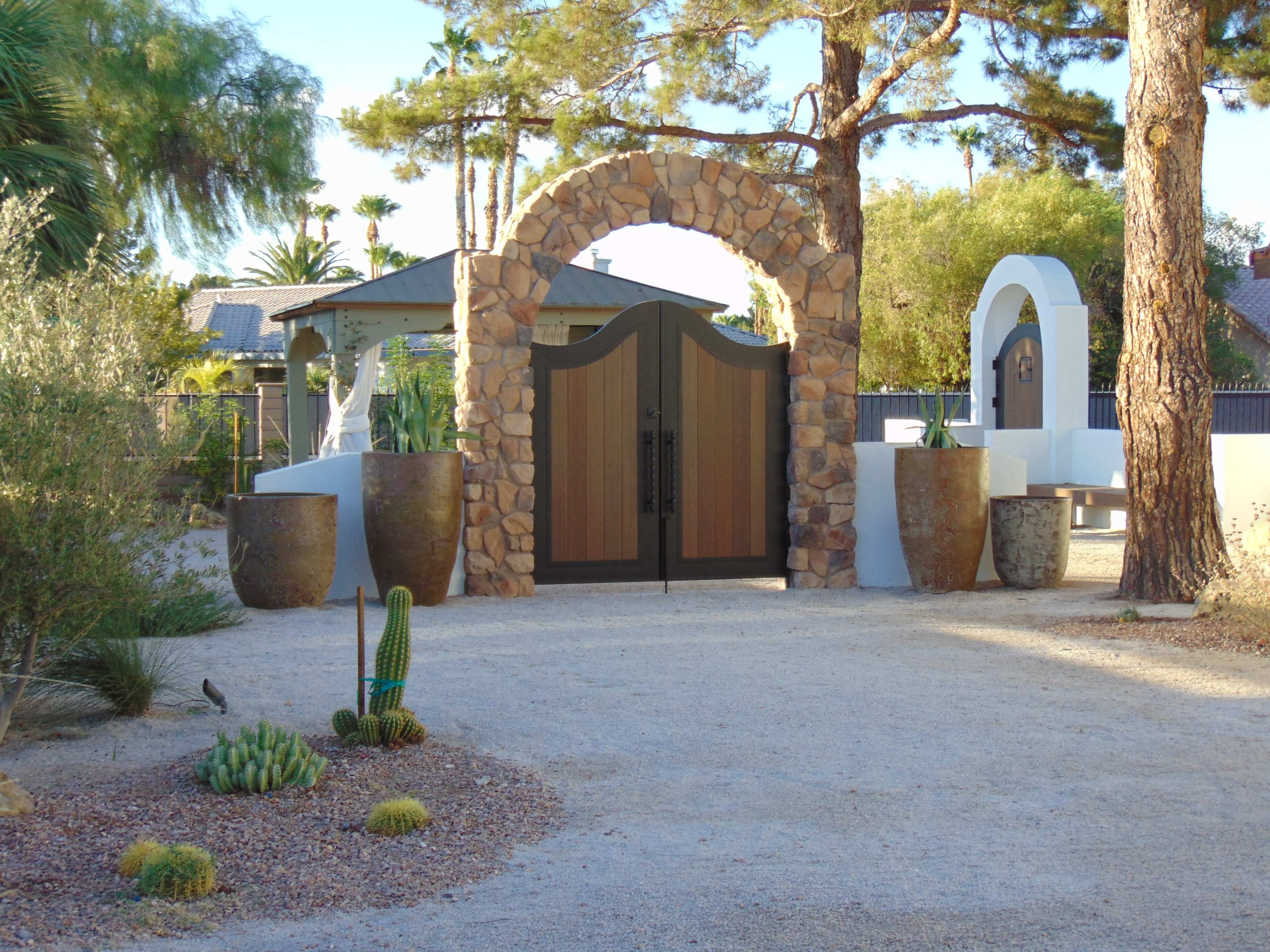 A stone archway with a wooden gate stands at the entrance to a landscaped yard featuring potted plants and gravel pathways.