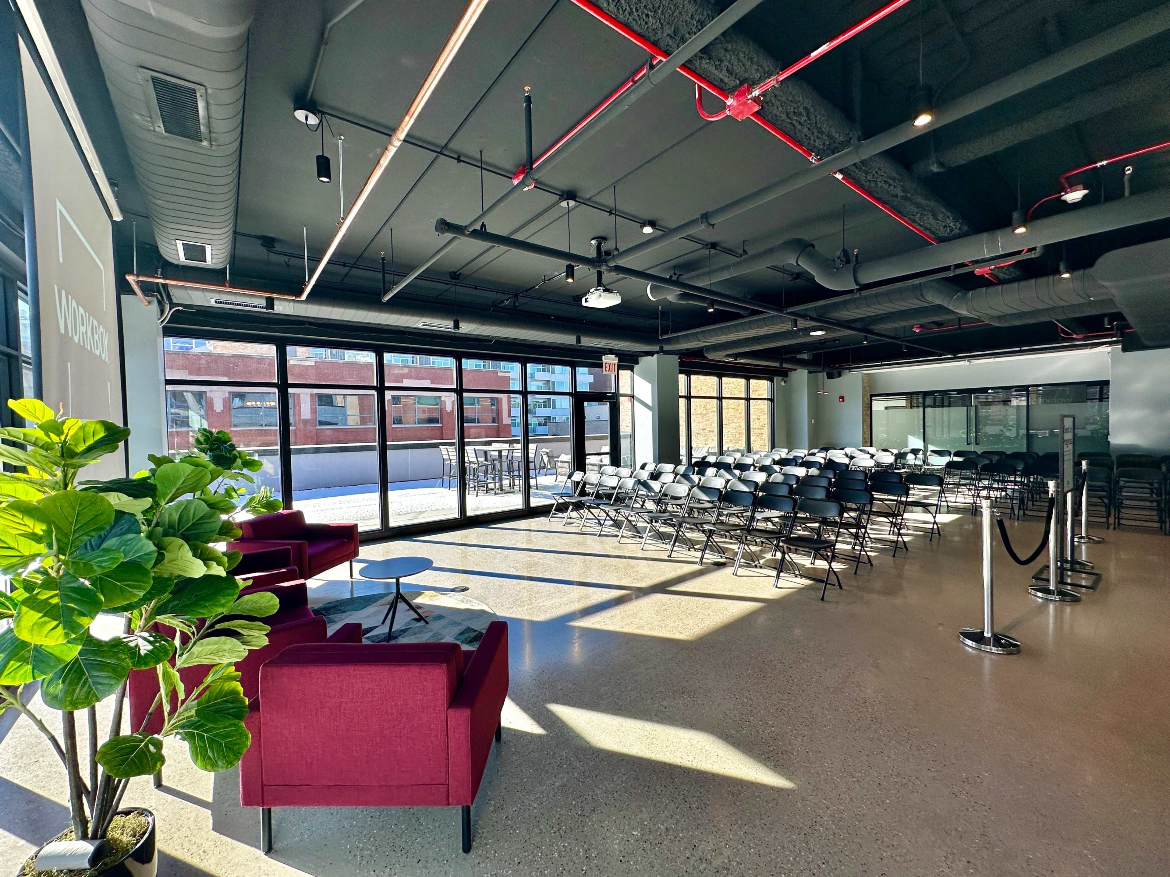 The image shows a modern event space with rows of chairs set up facing large windows and a green plant in the foreground.