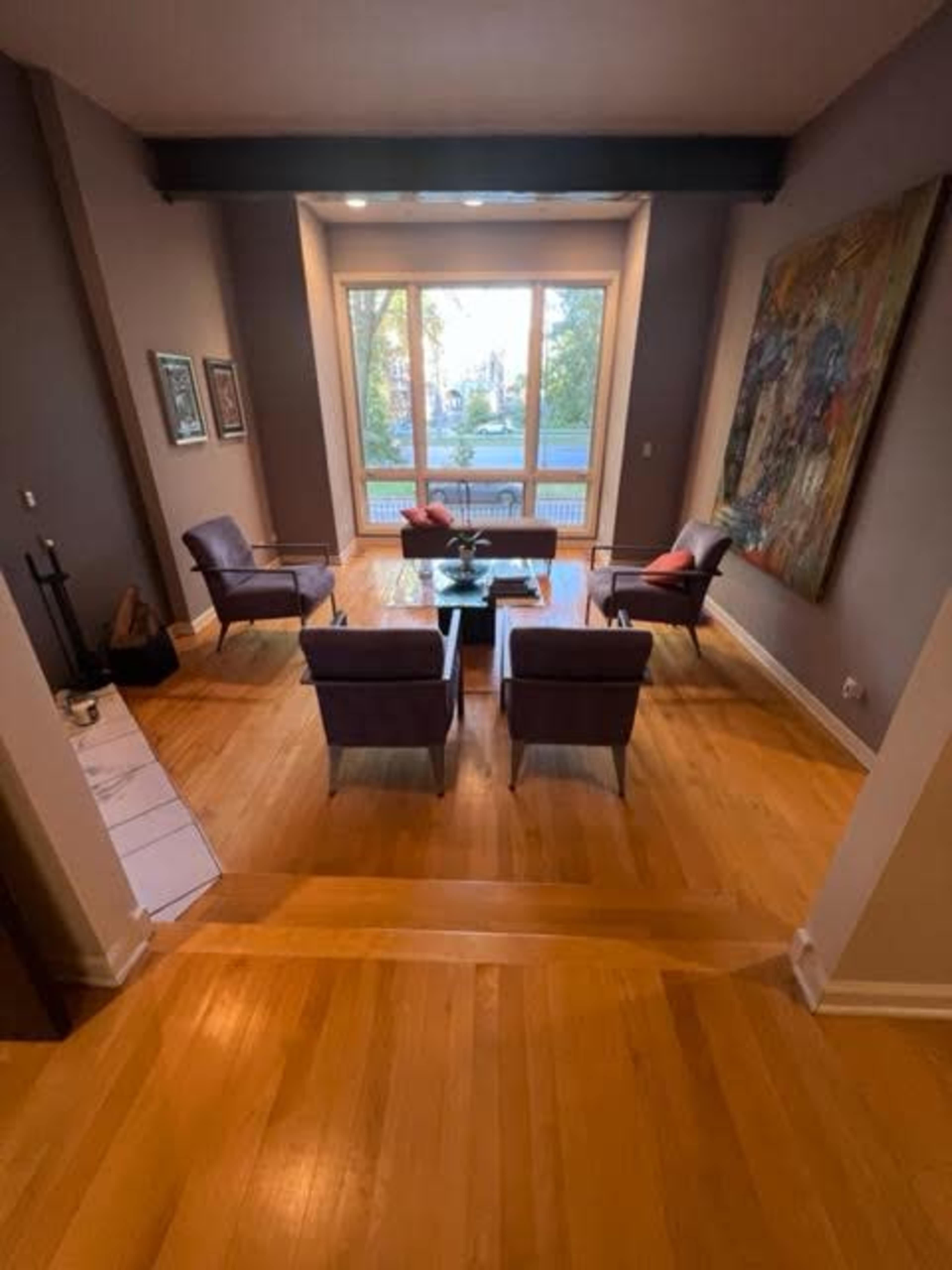 A spacious living area with hardwood floors, a central coffee table, and four chairs arranged around it, all set in front of large windows that provide natural light.