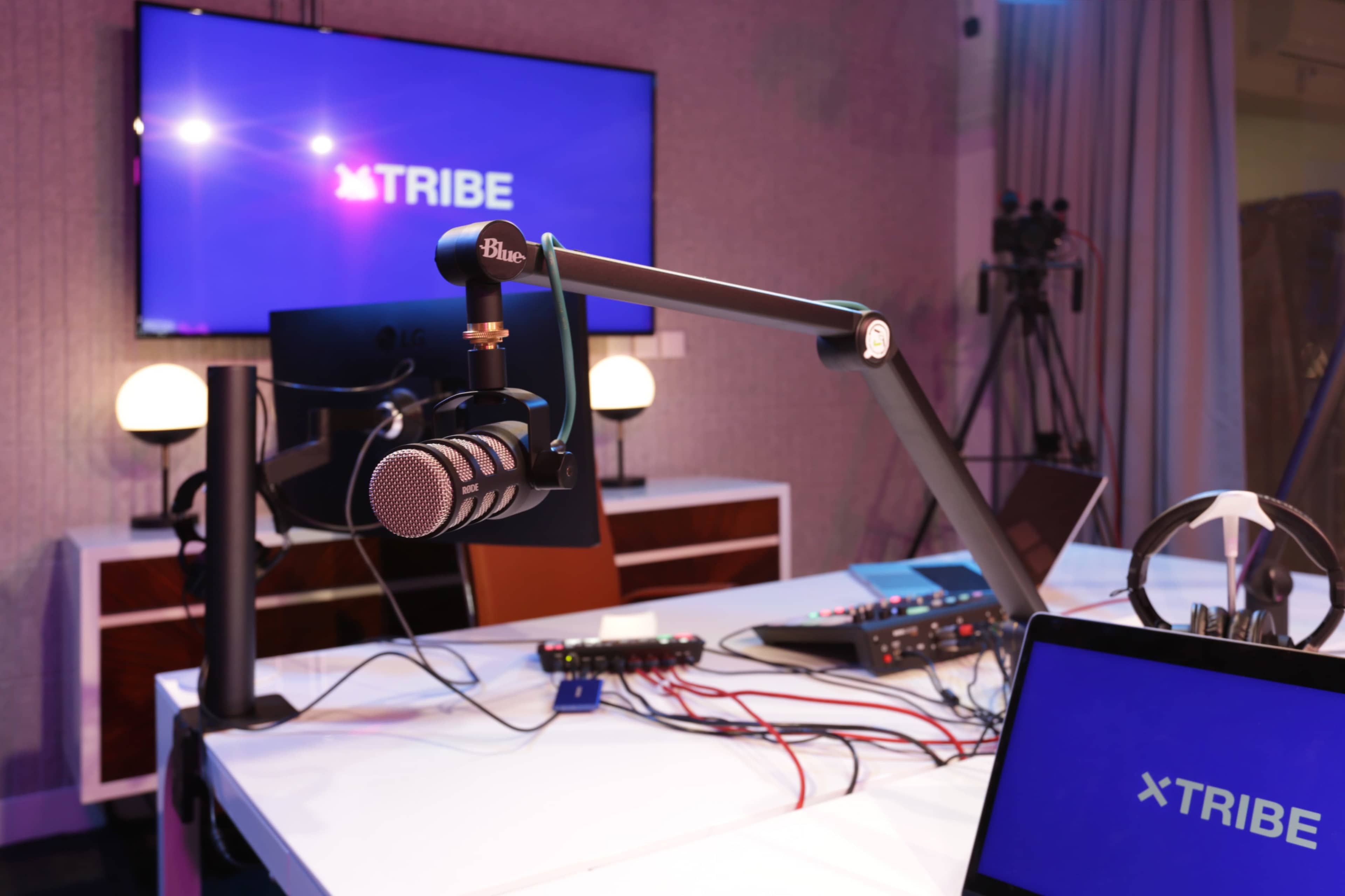 The image shows a modern podcasting studio featuring a microphone on an adjustable arm, a digital mixing console, and a large screen displaying the word "TRIBE."