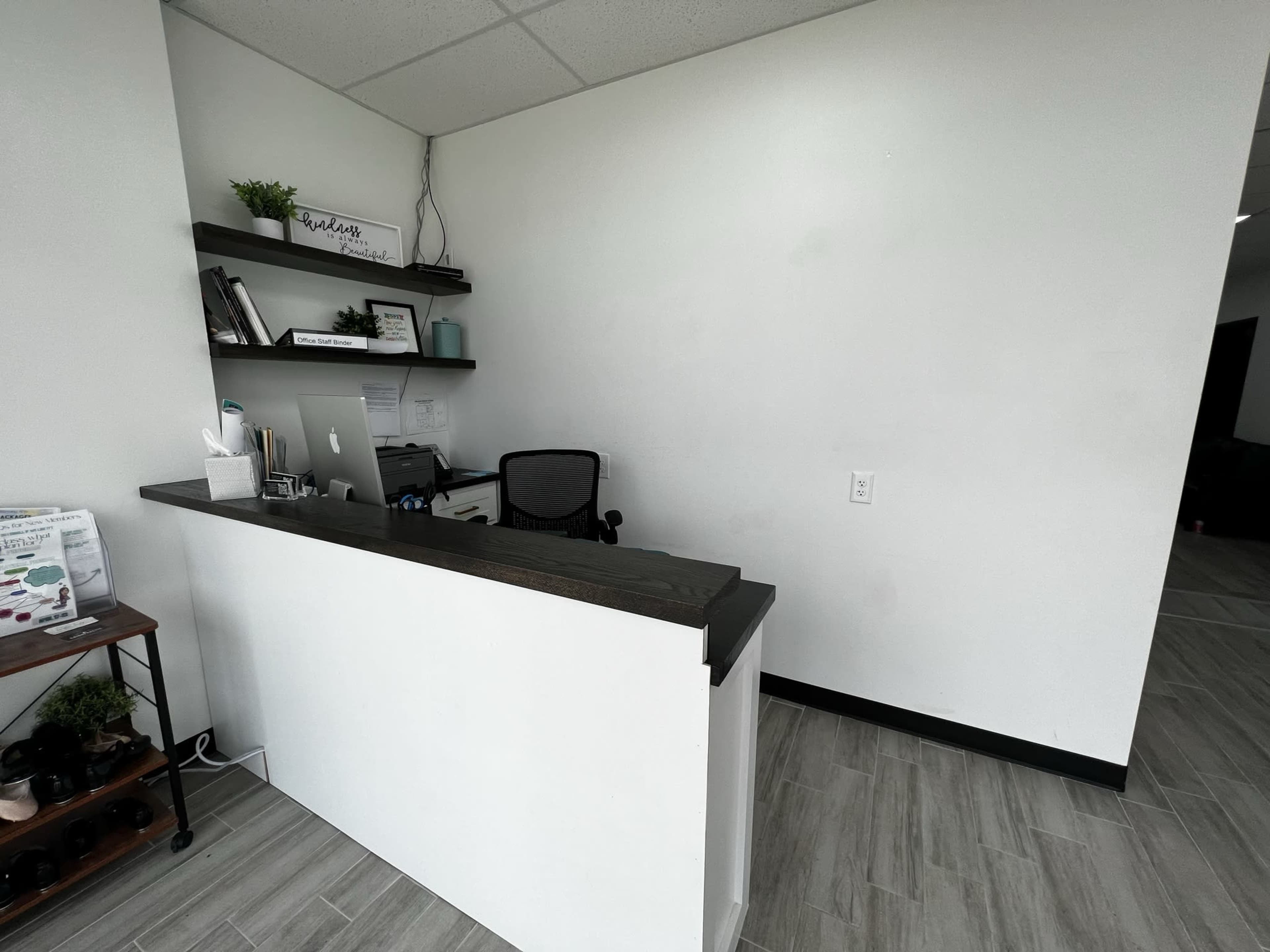 A minimalist reception desk with a computer and books on a shelf against a white wall.