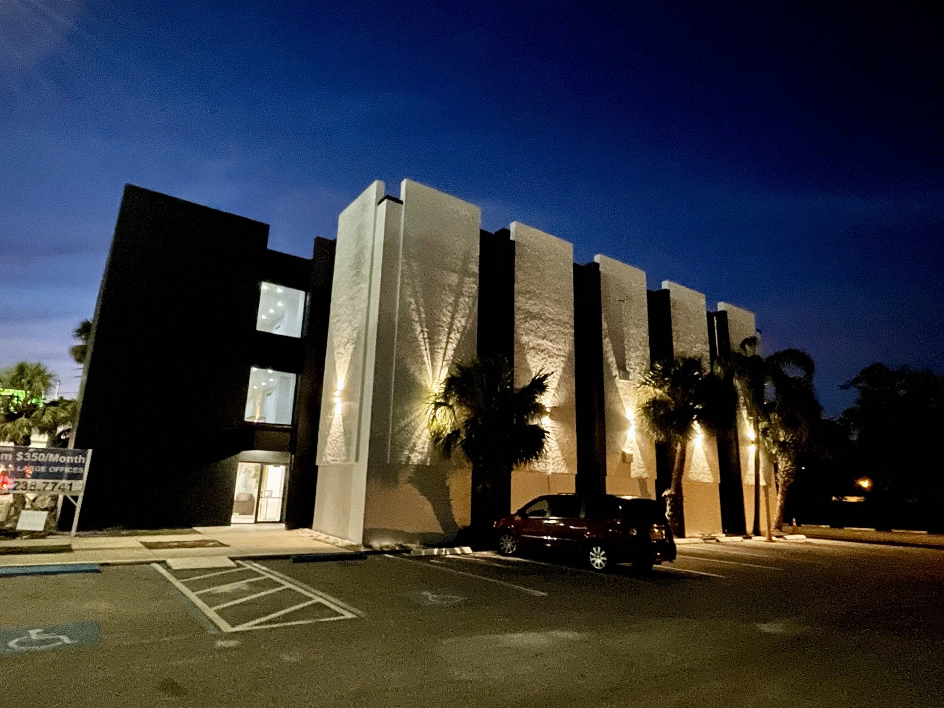 A modern building with a textured exterior stands in a parking lot at dusk, illuminated by lights on the façade.
