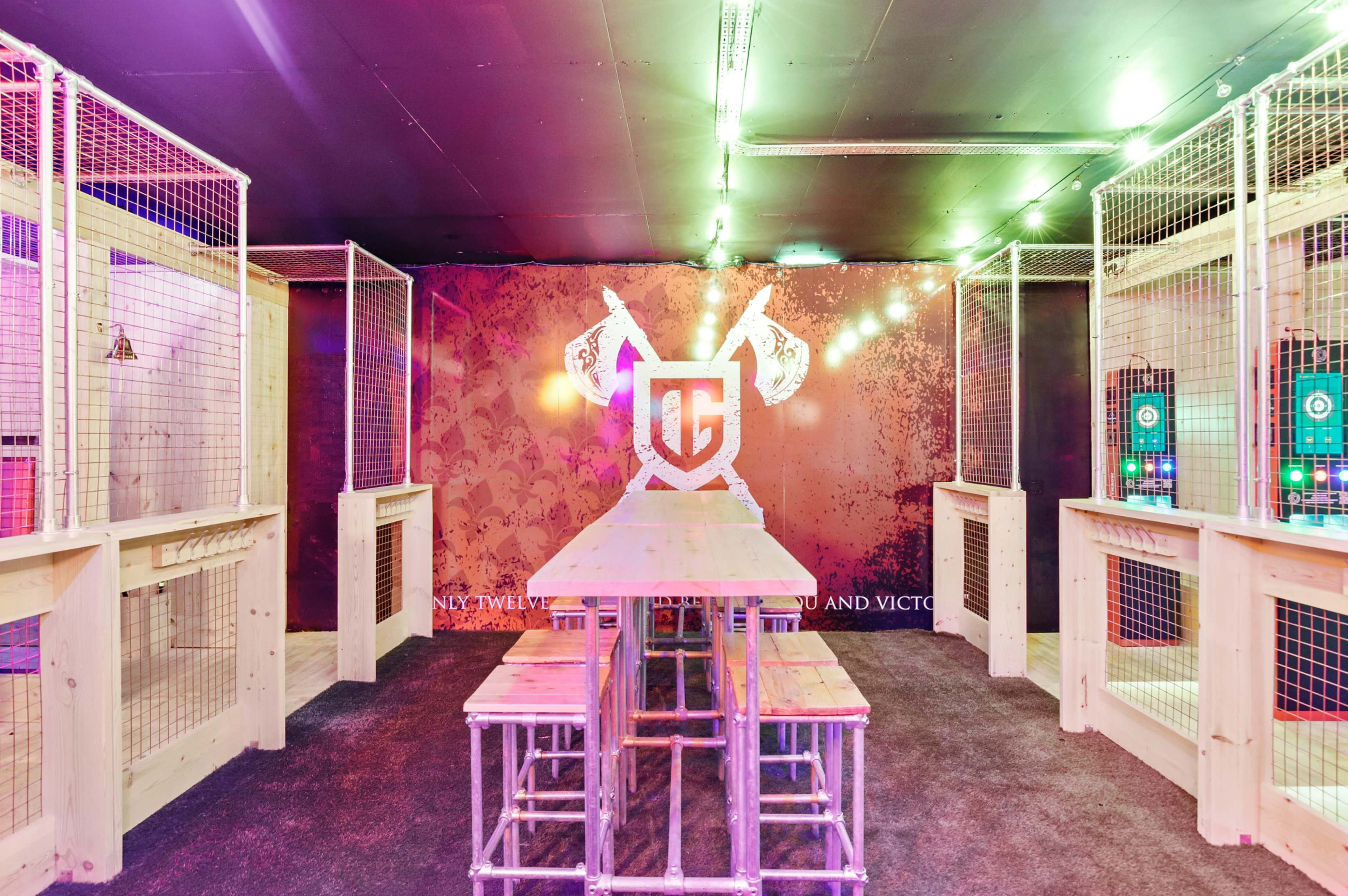 An indoor axe-throwing venue featuring wooden tables and enclosure fencing, illuminated by colorful lights.