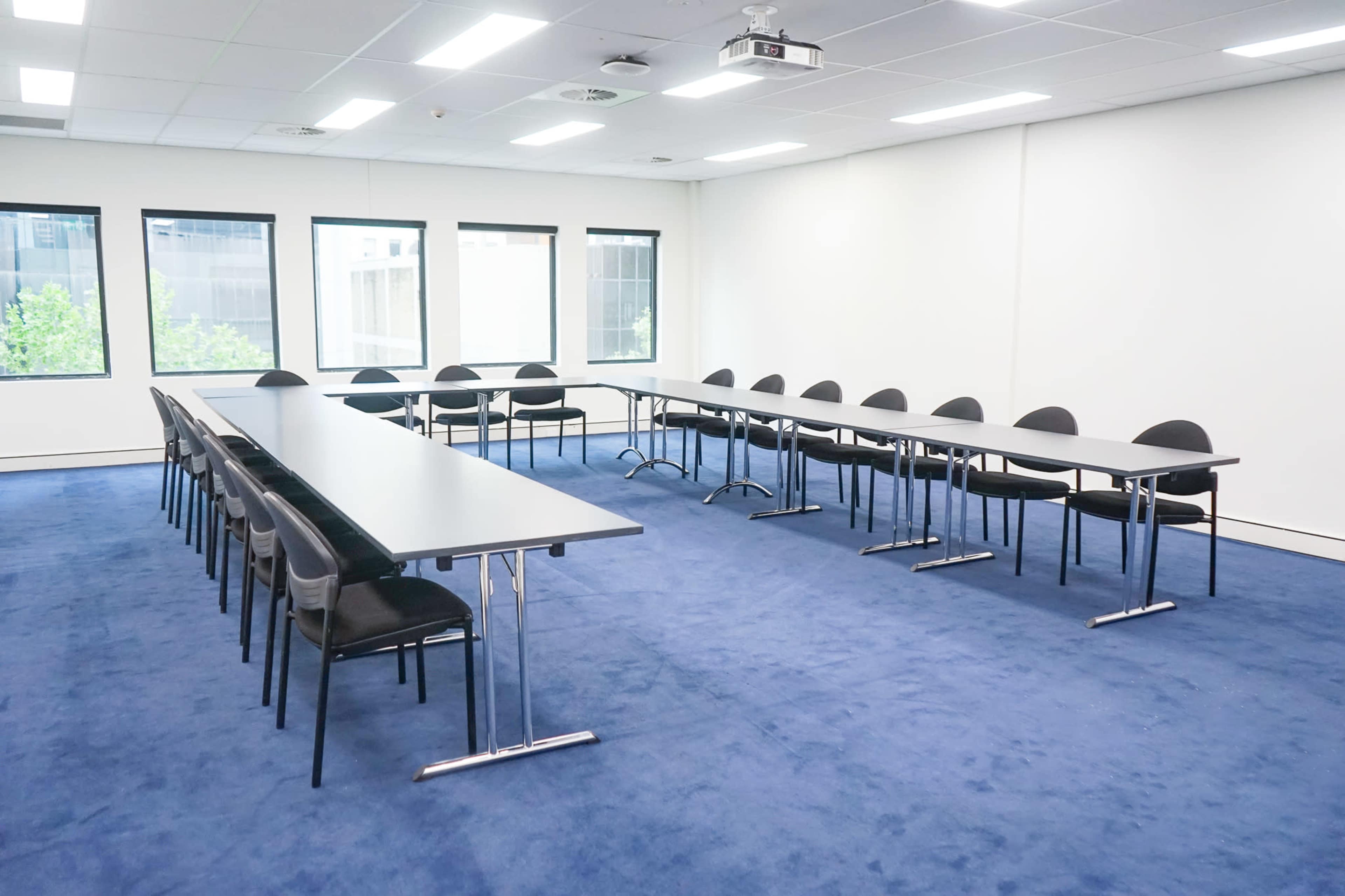 A spacious conference room with two long tables arranged in a U-shape and multiple black chairs on a blue carpet.
