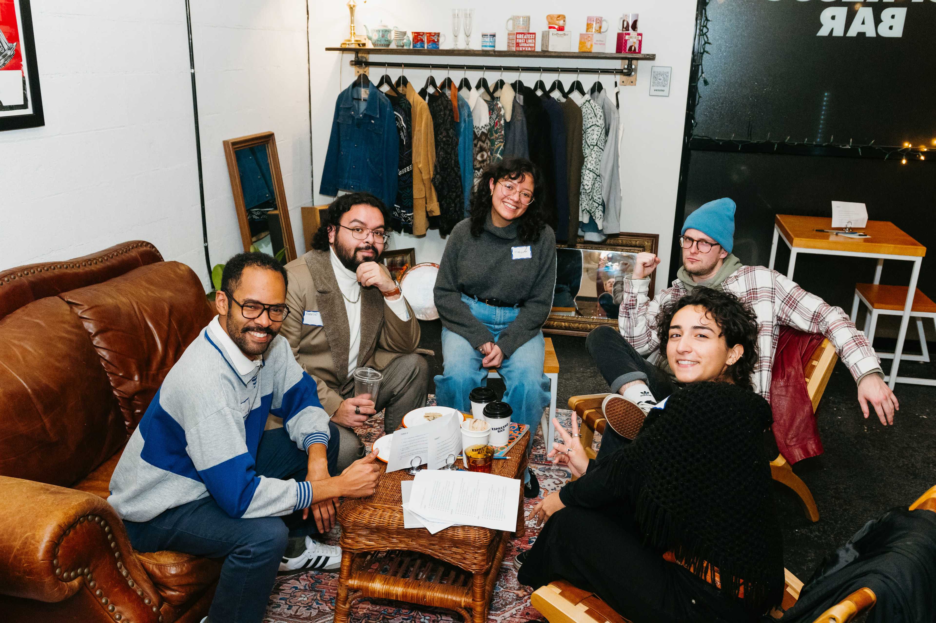 A group of five people sits in a cozy, casual lounge area with clothing displayed in the background.