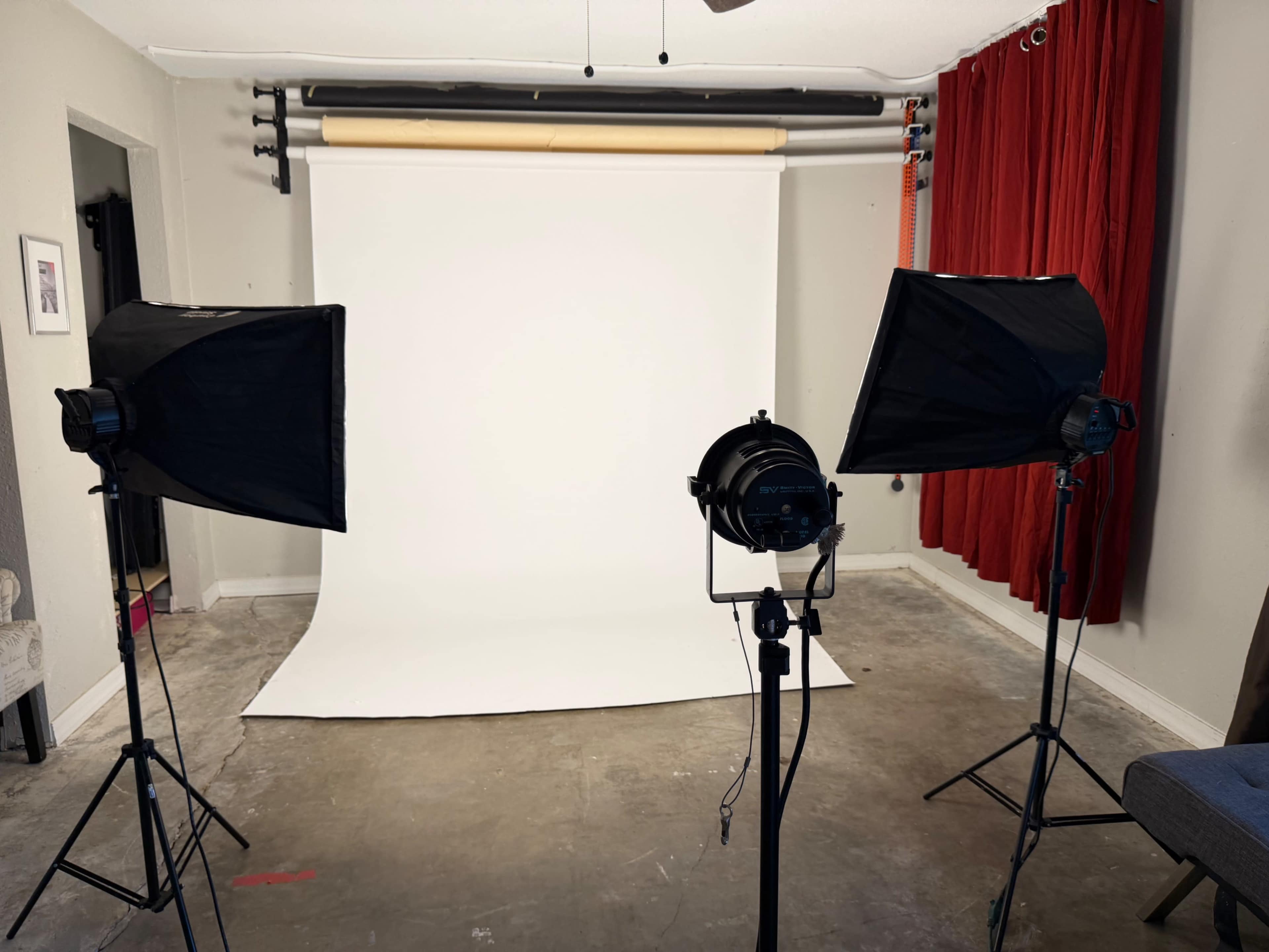 The image shows a photography studio with a white backdrop and three lighting setups positioned symmetrically in front of it.