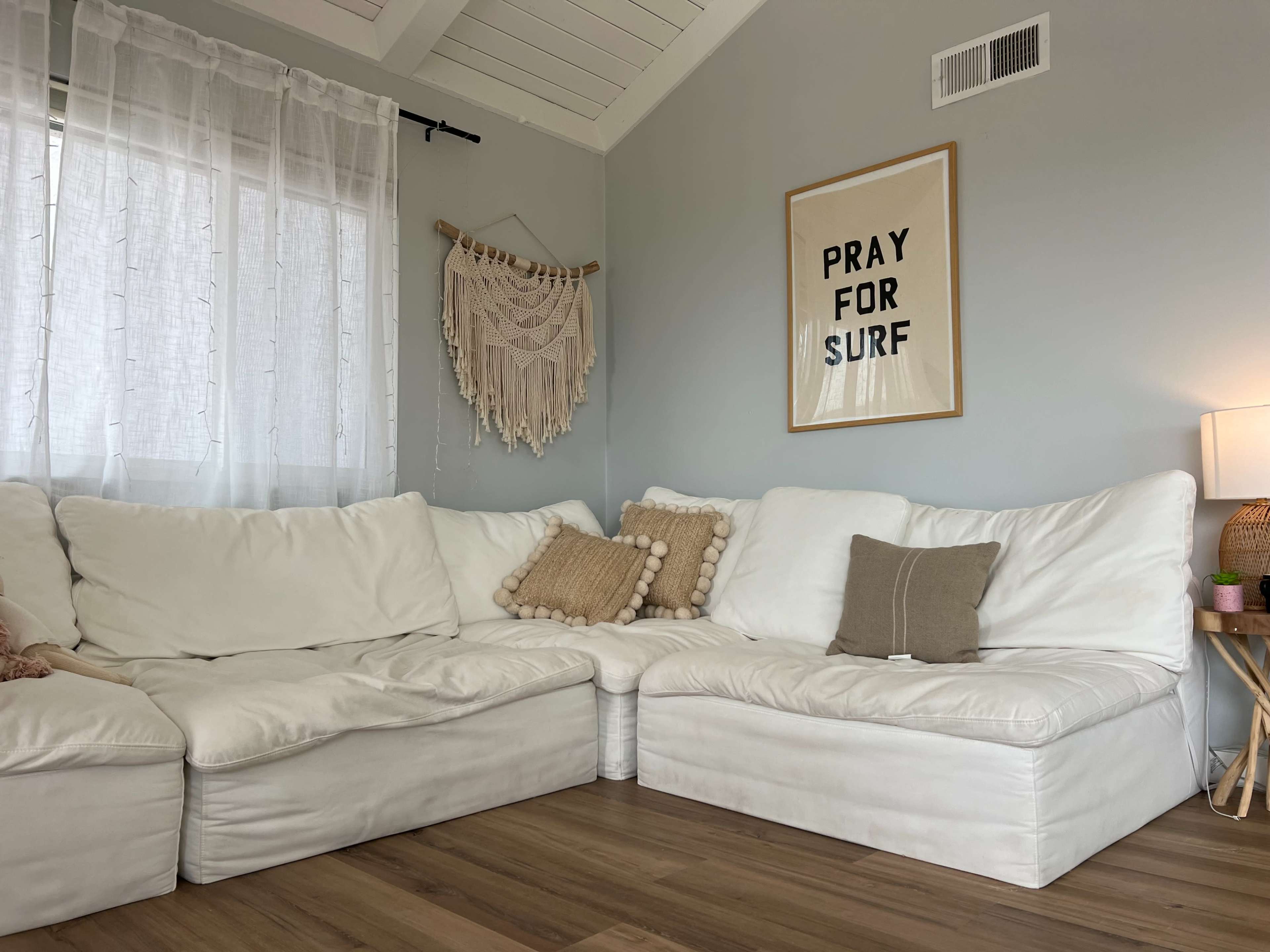 A white sectional sofa with decorative pillows is positioned in a light-filled corner of a room featuring a macramé wall hanging and a framed motivational poster.