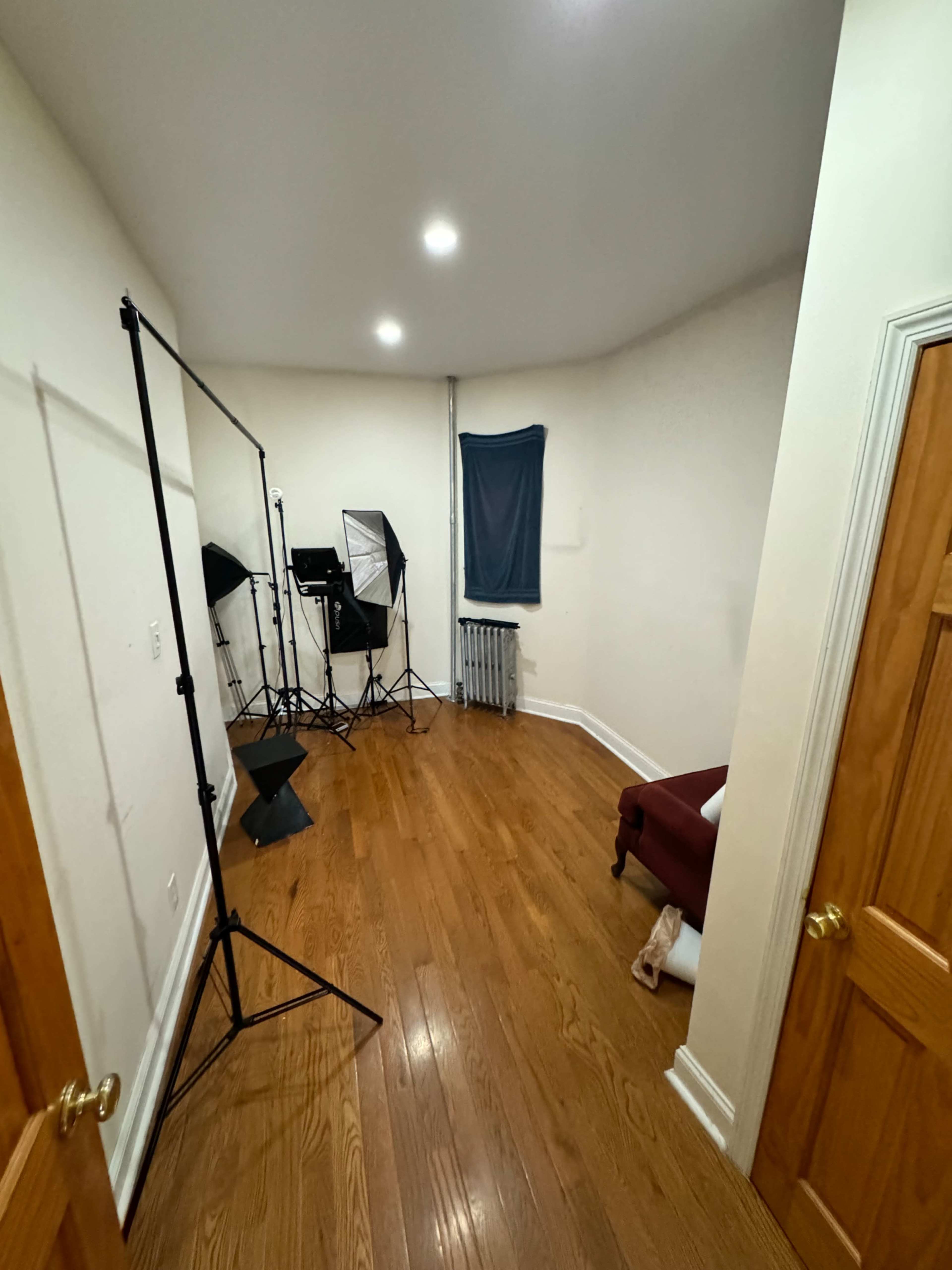 The image shows a small, well-lit room with wooden flooring, featuring photography equipment, a blue curtain on a window, and a maroon chair.