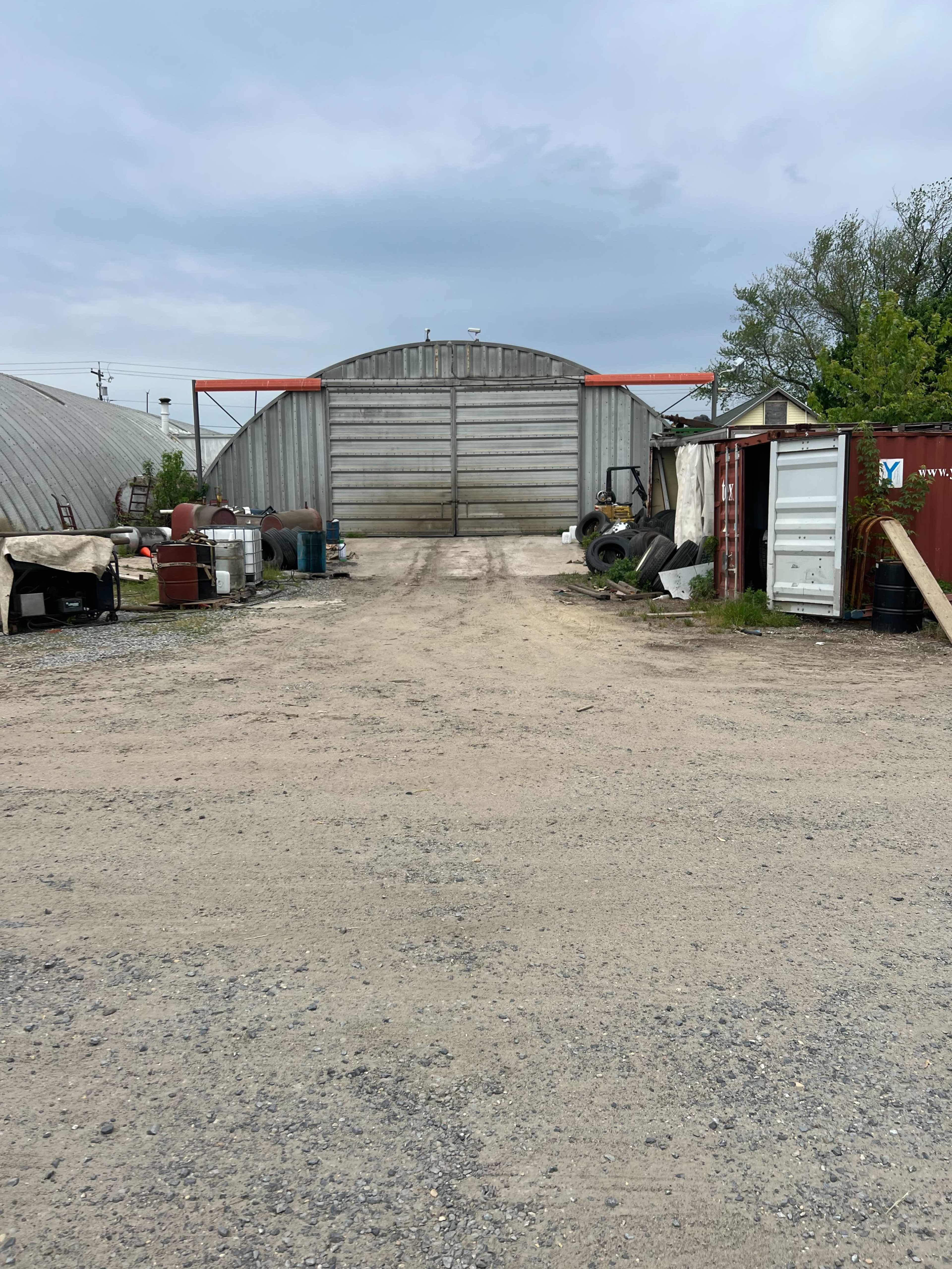 The image shows a gravel road leading to a large, metal barn-like structure with a sliding door, flanked by containers and scattered equipment.