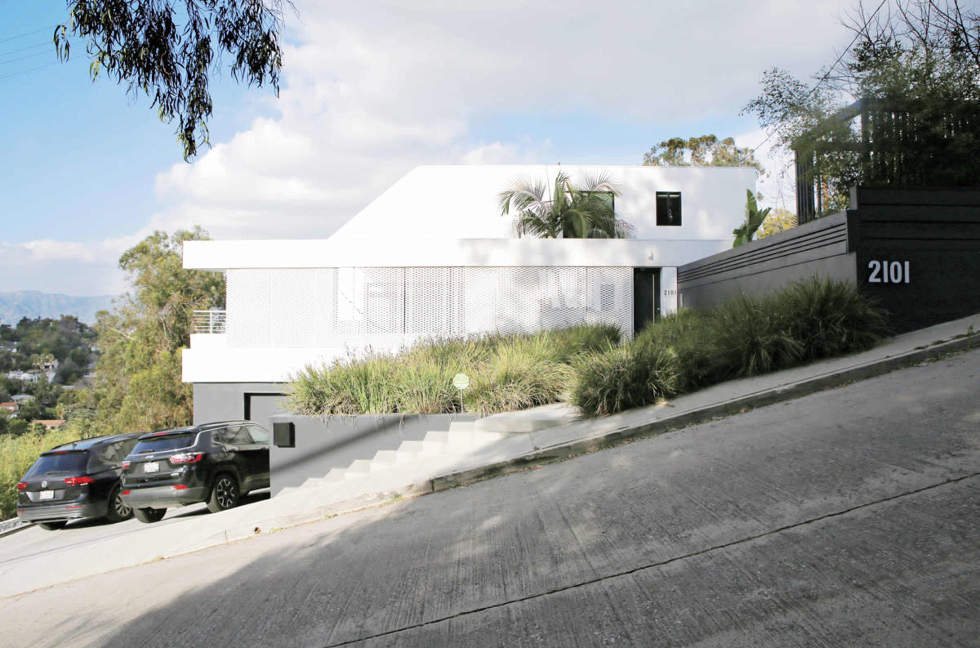 A modern white house with large windows is situated on a sloped driveway, flanked by two parked vehicles and surrounded by greenery.