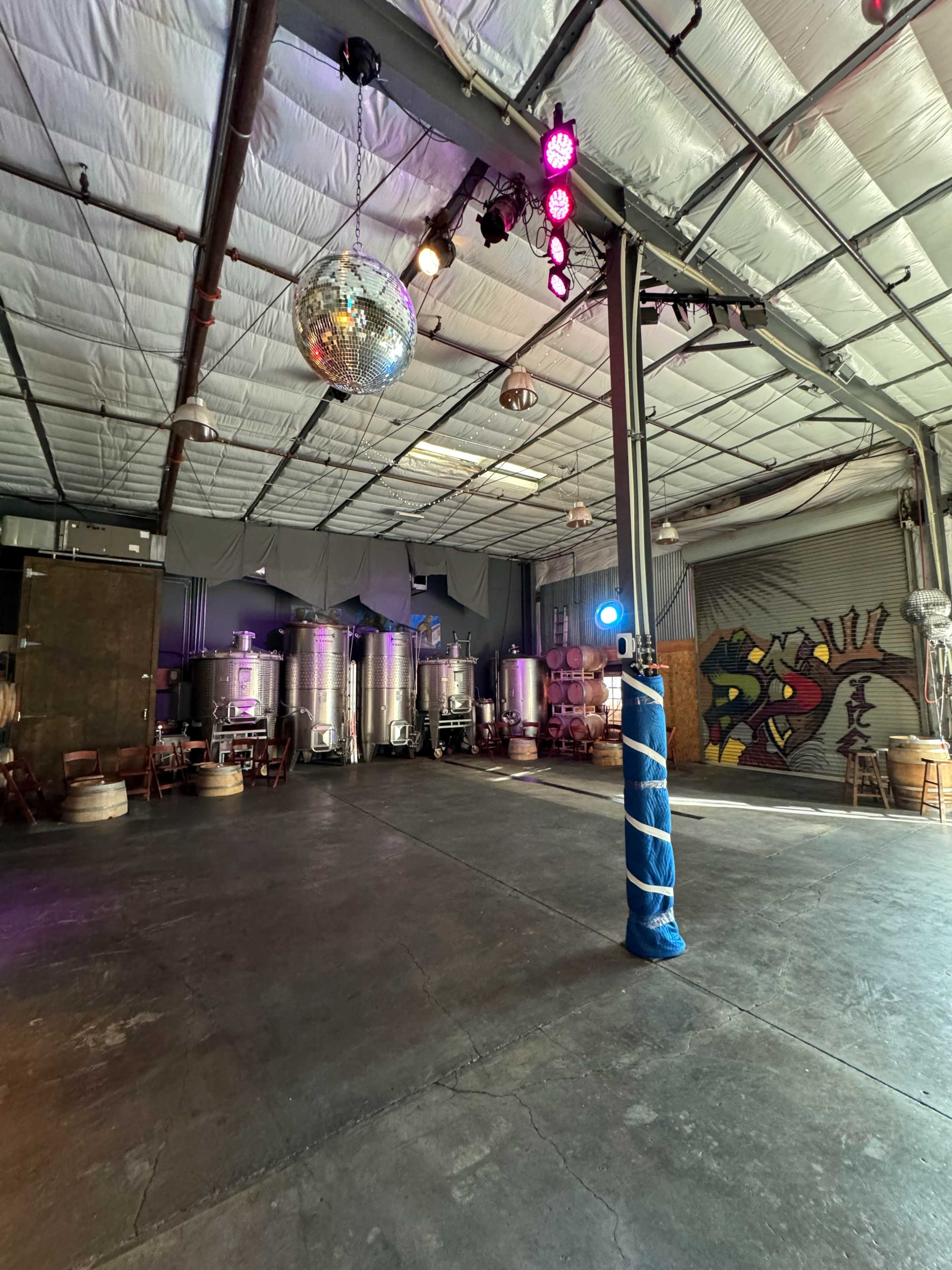 An industrial space with metal brewing tanks, a disco ball hanging from the ceiling, and colorful stage lights illuminating a concrete floor.