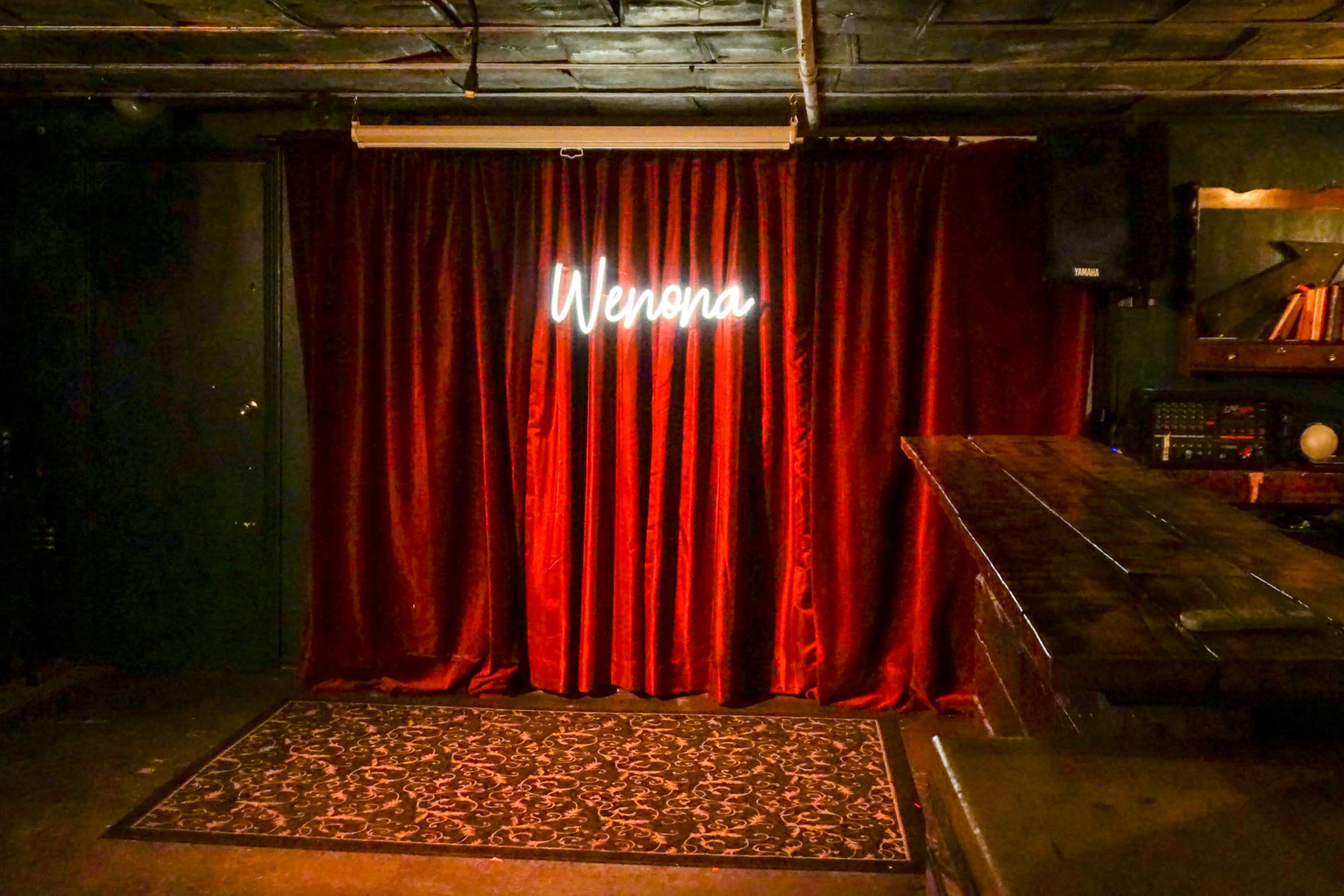 A dimly lit stage with red velvet curtains and a neon sign that reads "Wenona."