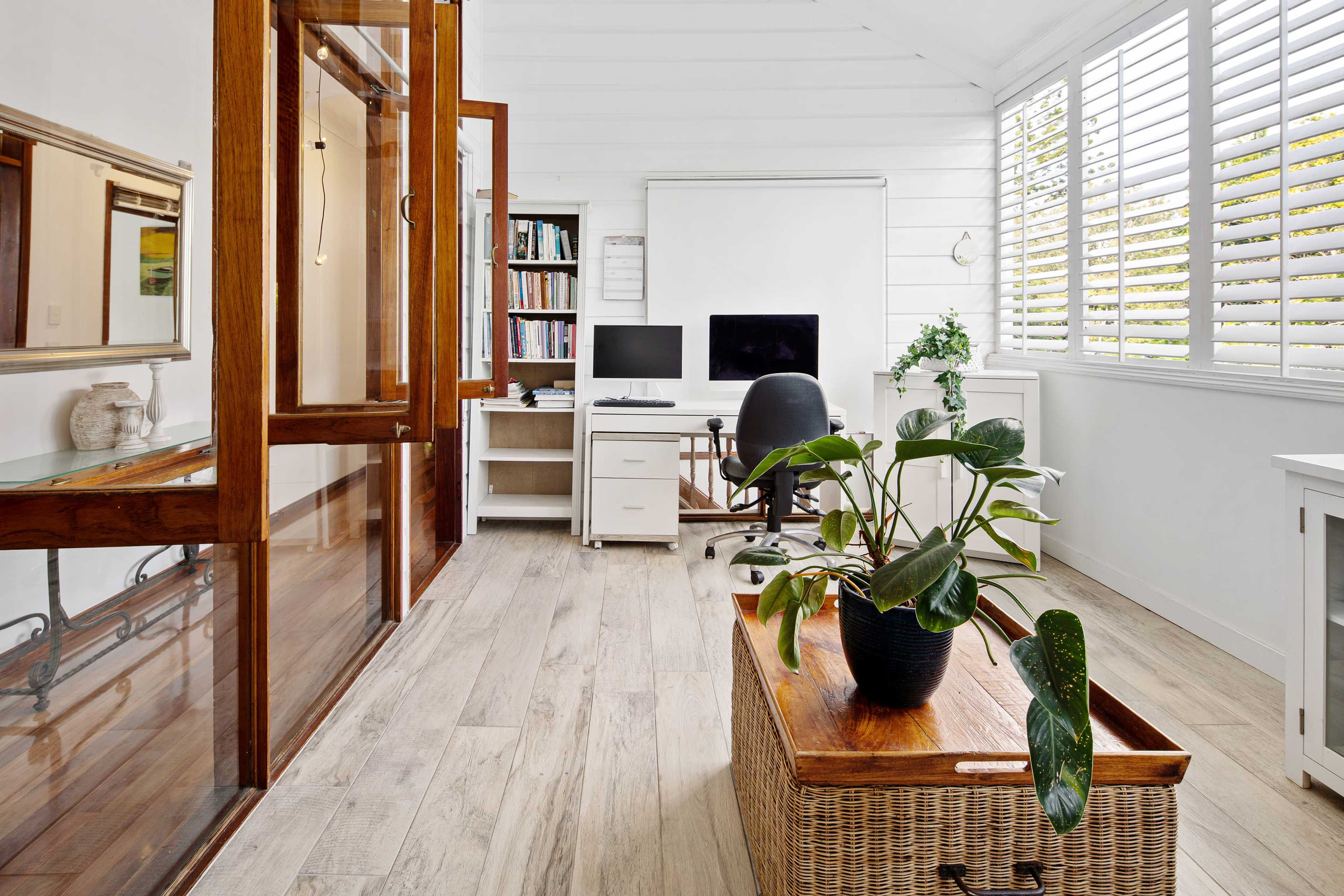 The image shows a home office with a wooden desk, a computer setup, a shelf filled with books, and a potted plant on a wicker storage chest.