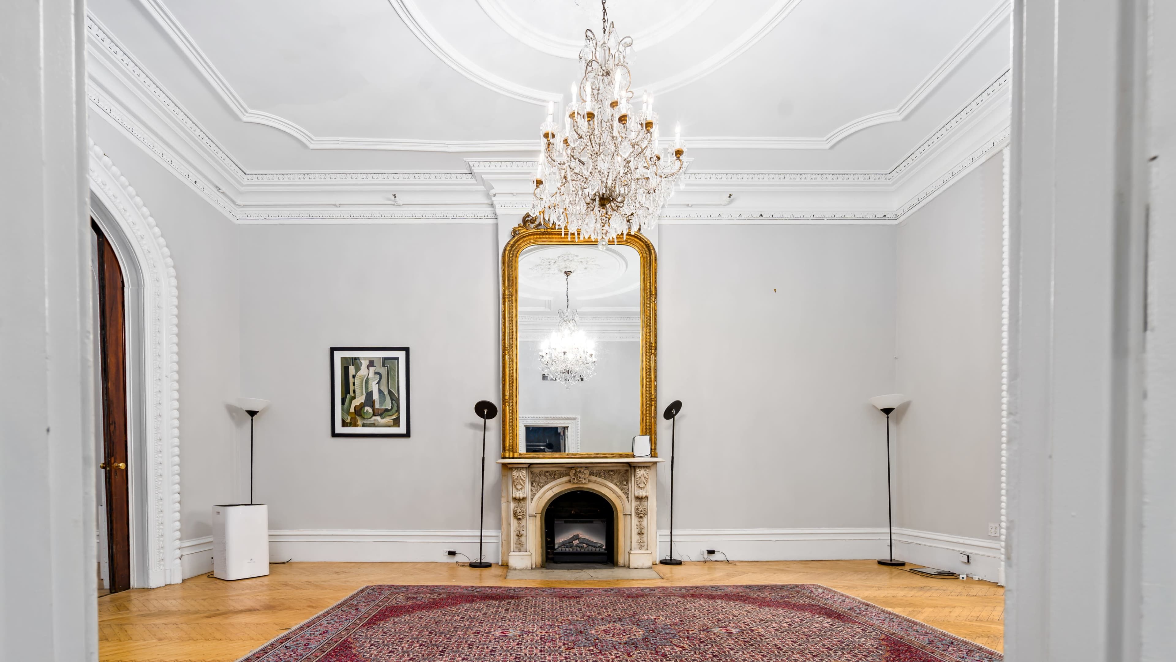 The room features a chandelier, a large gilded mirror above a decorative fireplace, and two standing lamps positioned near the walls.