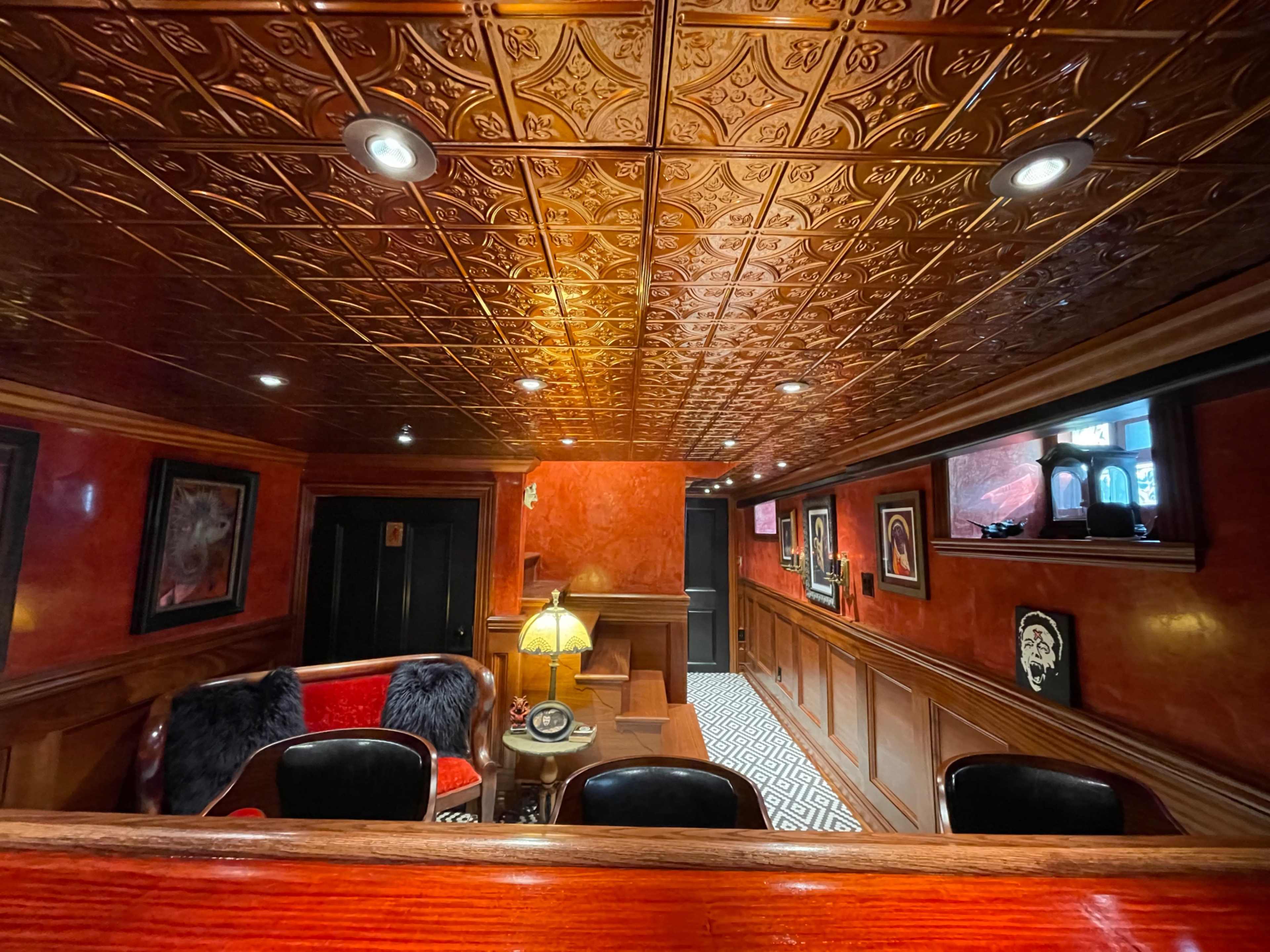 The image depicts a cozy basement room featuring a red and black color scheme, ornate ceiling tiles, and plush seating arrangements.