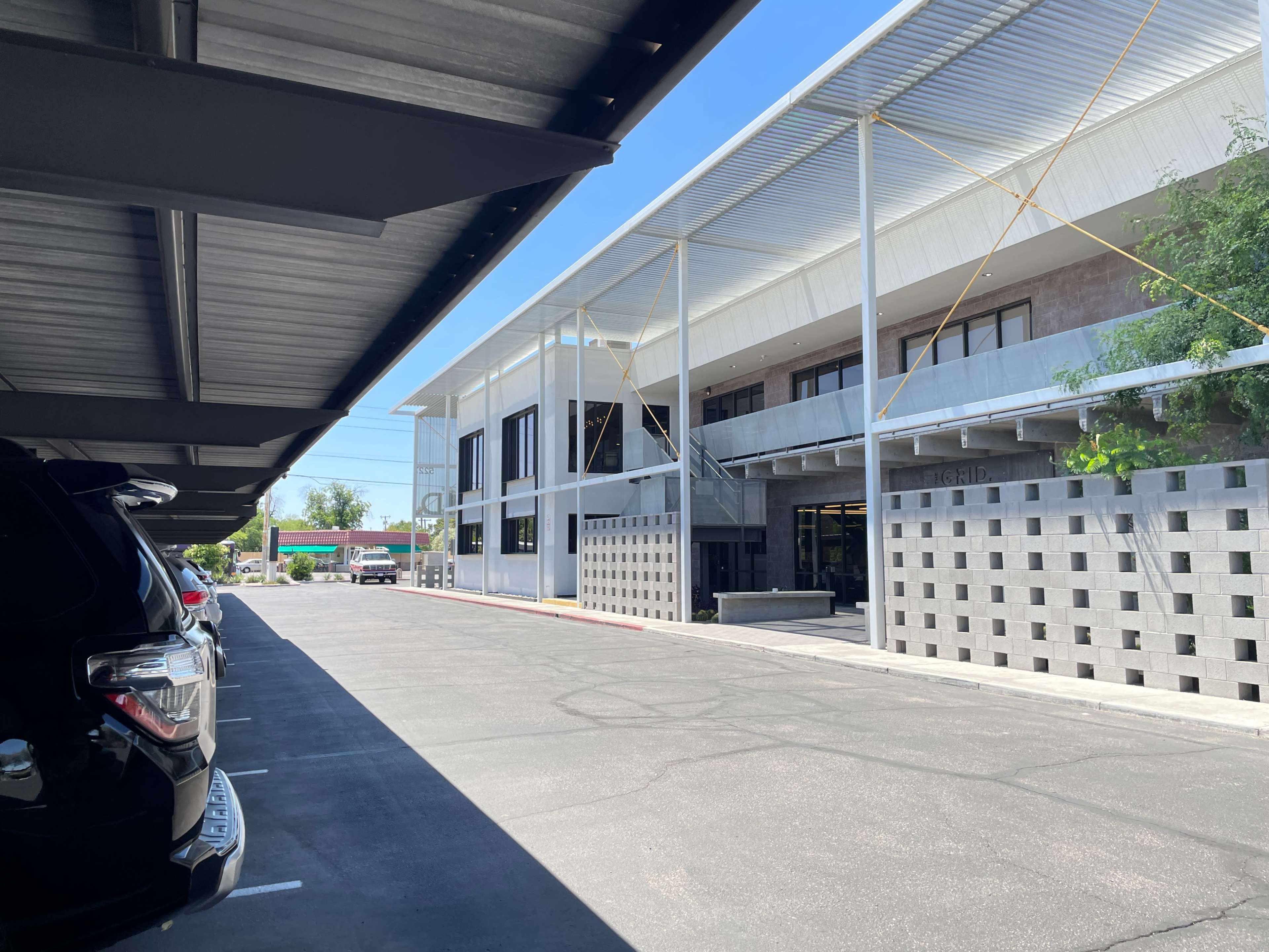 The image shows a modern building with a covered parking area in front, featuring a minimalist architectural design and a clear blue sky above.