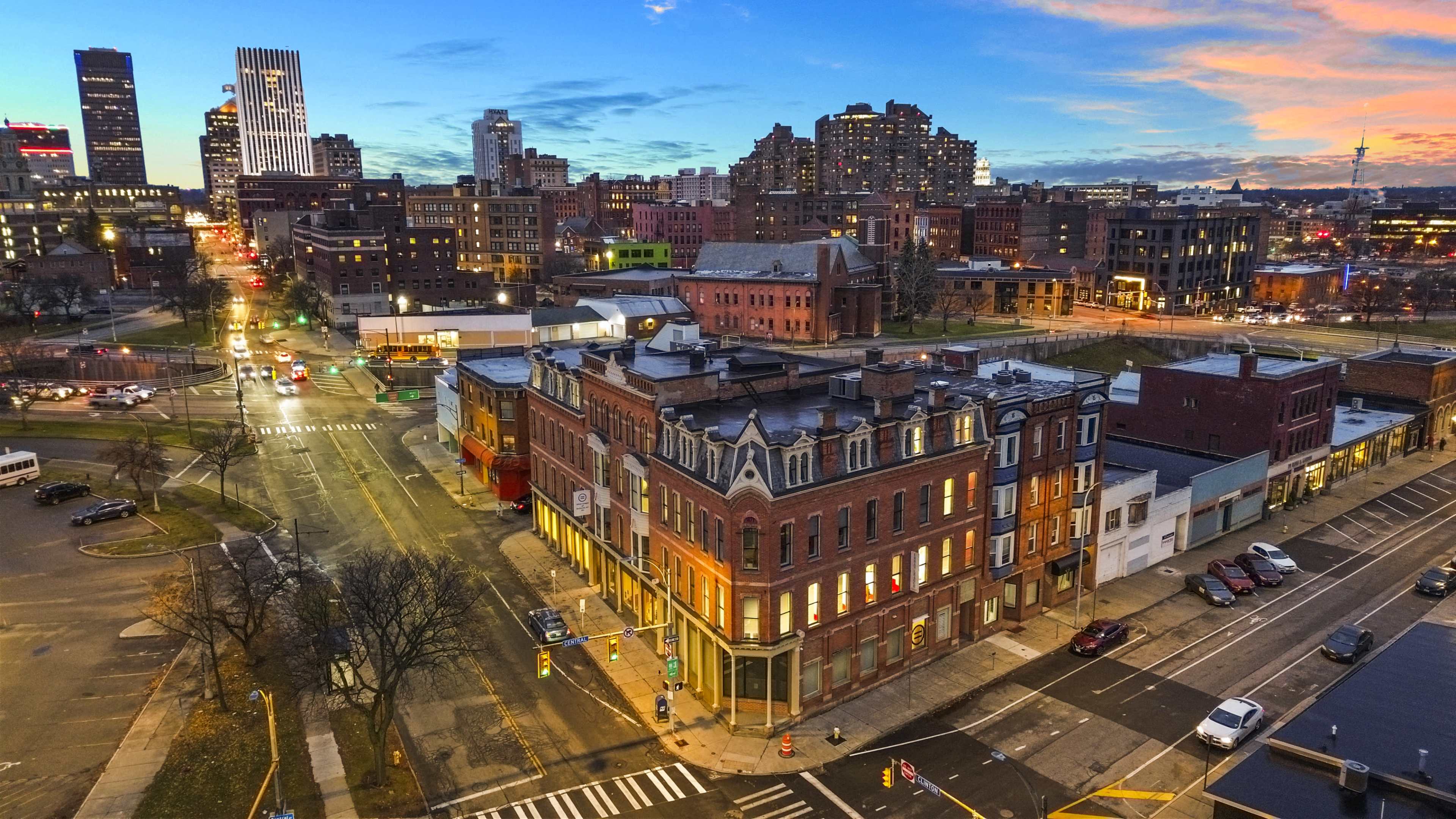 A vibrant cityscape at dusk, featuring a historic building at a street intersection with modern skyscrapers in the background.