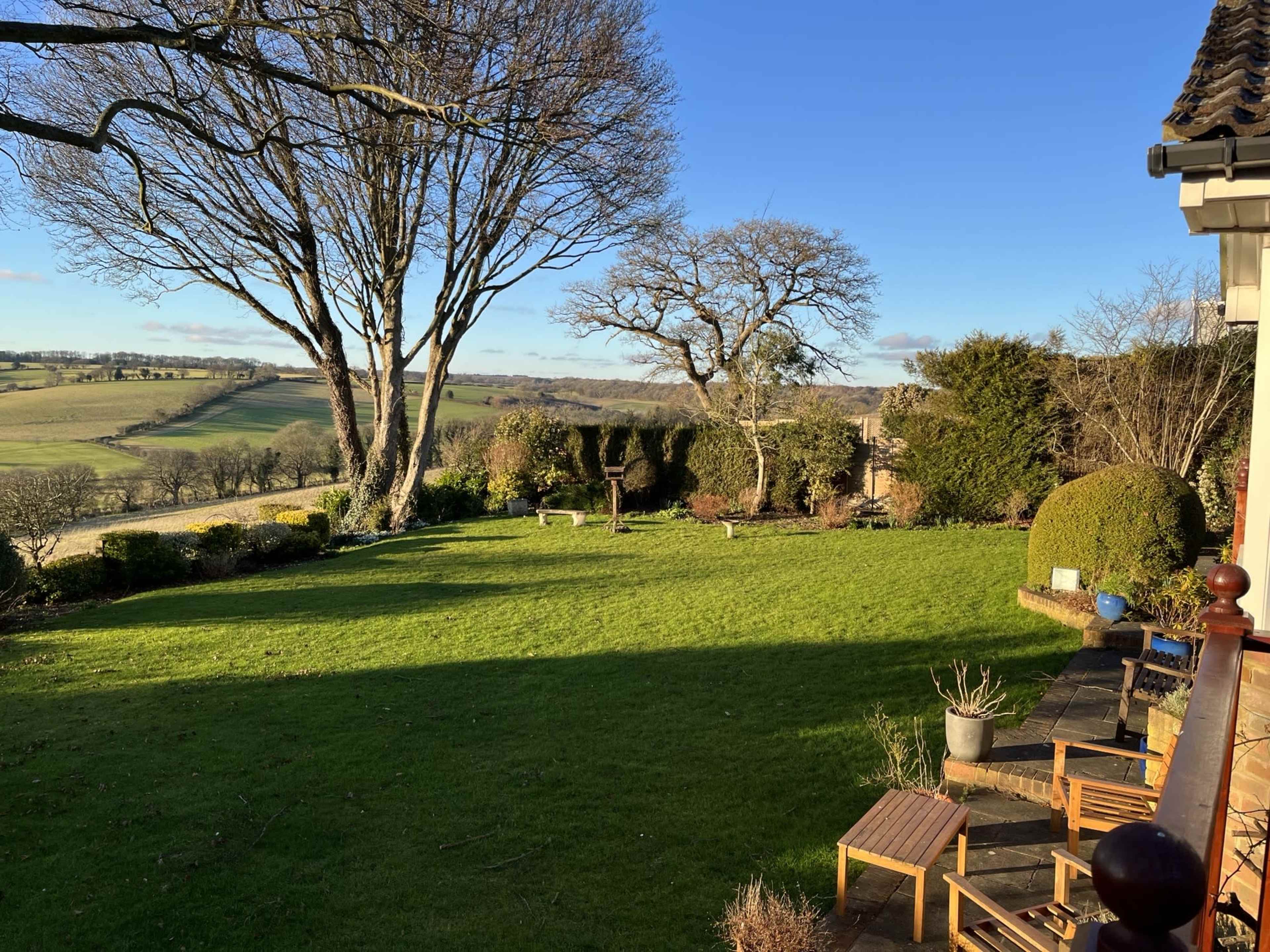 A well-maintained garden with neatly trimmed grass, a few trees, and patio furniture overlooking rolling hills.