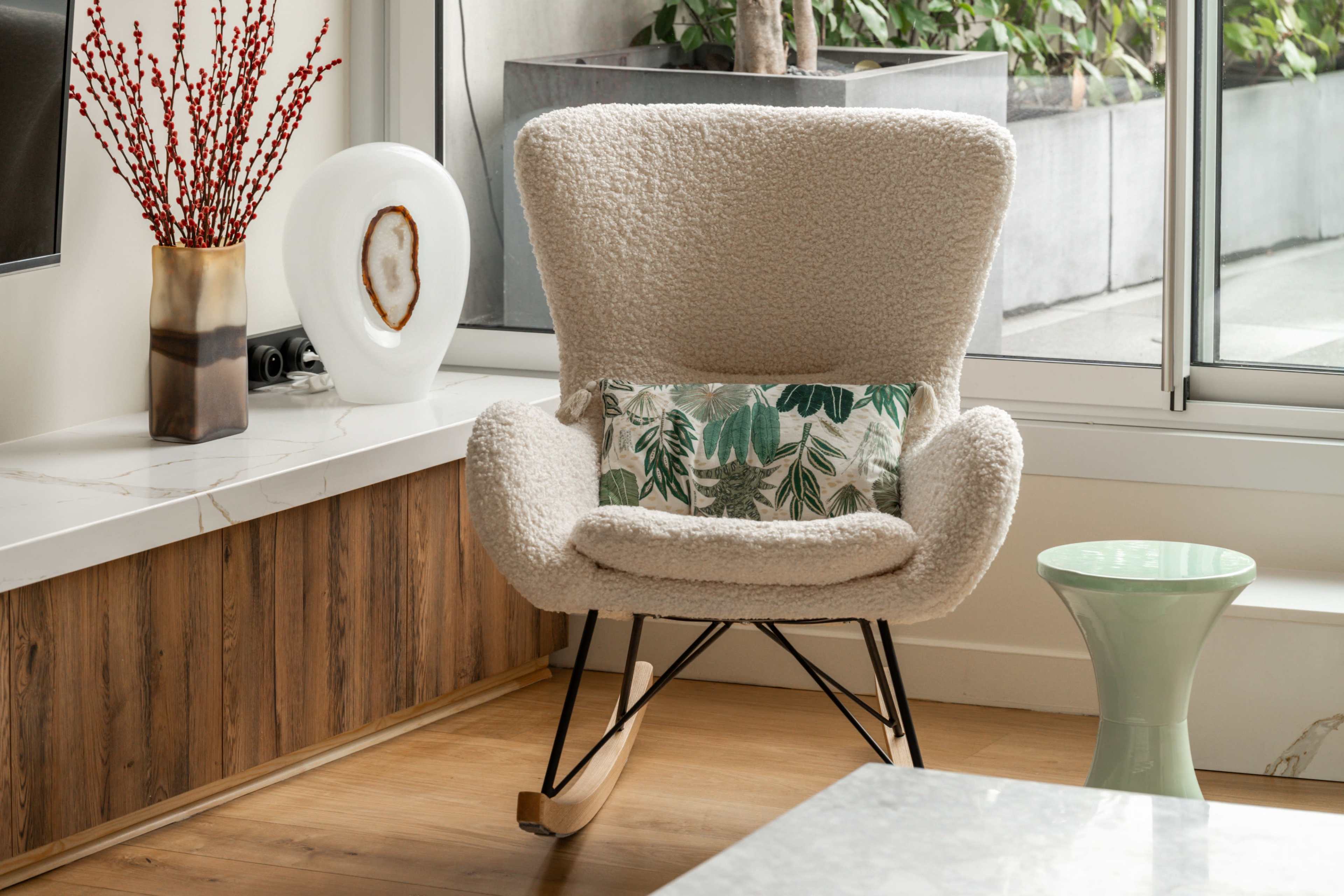 The image shows a cozy, modern living space featuring a textured rocking chair with a leafy patterned pillow, a small green side table, and decorative plants visible through a large window.