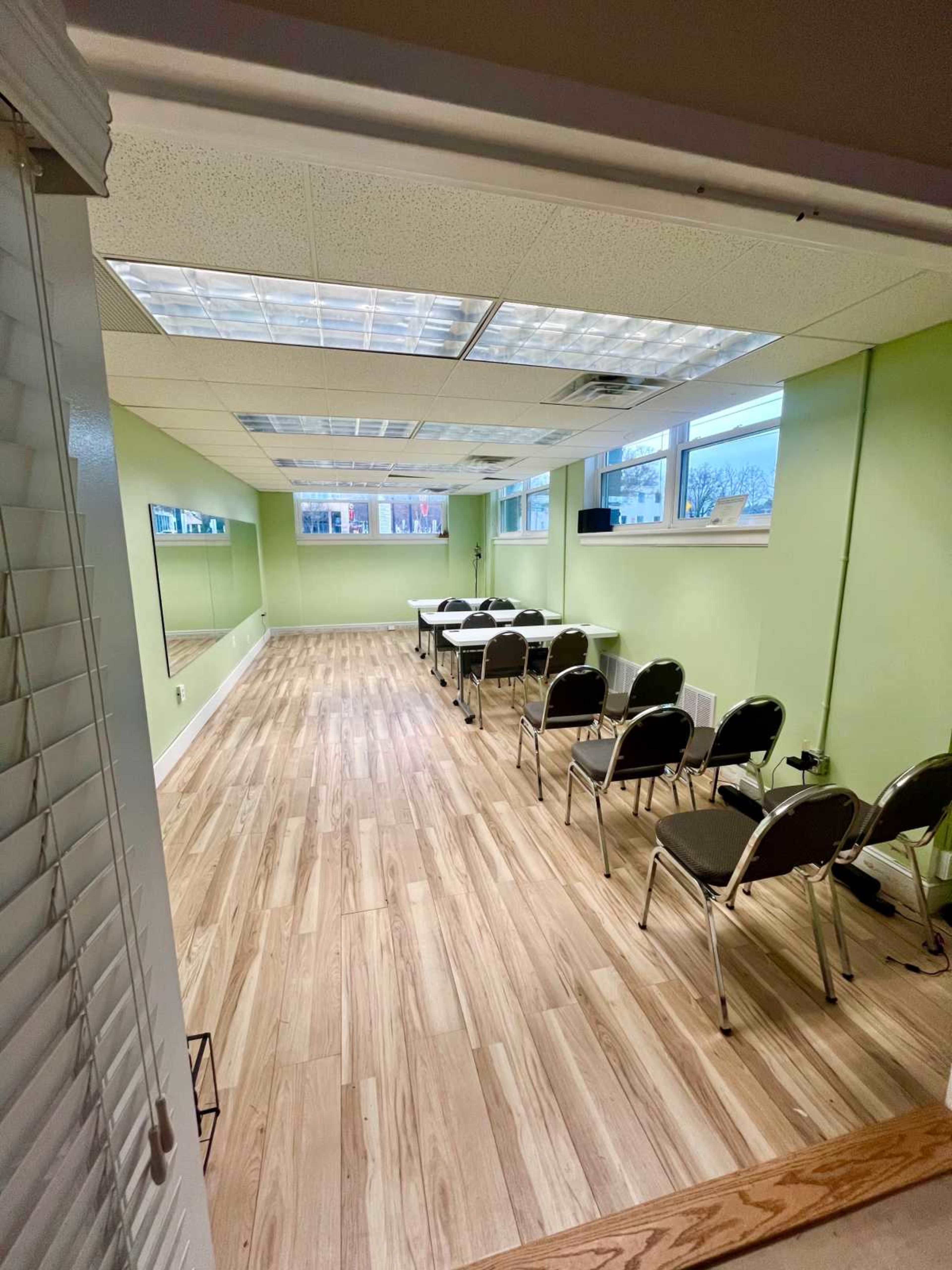The image shows a vacant room with light green walls, wooden flooring, and several chairs arranged in rows facing a table near the front.