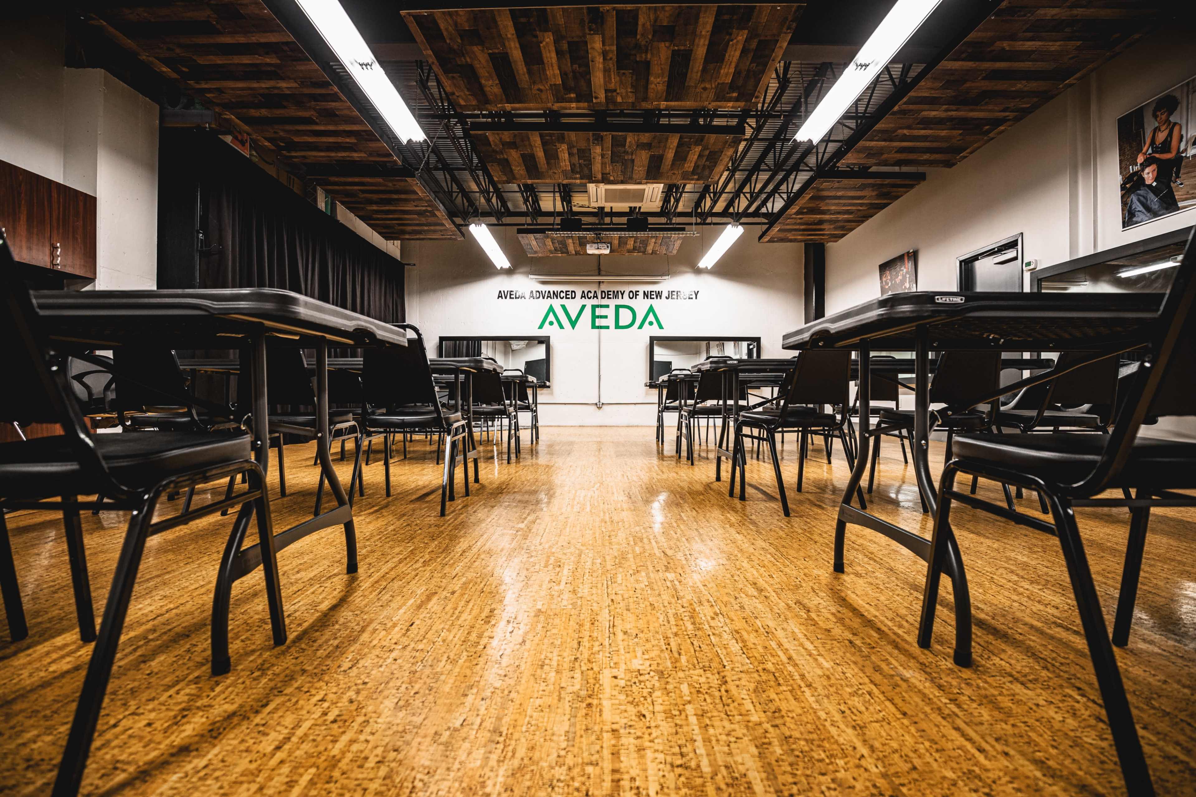 A classroom setup with empty black chairs and desks arranged in a line, featuring a wall marked with the Aveda logo.