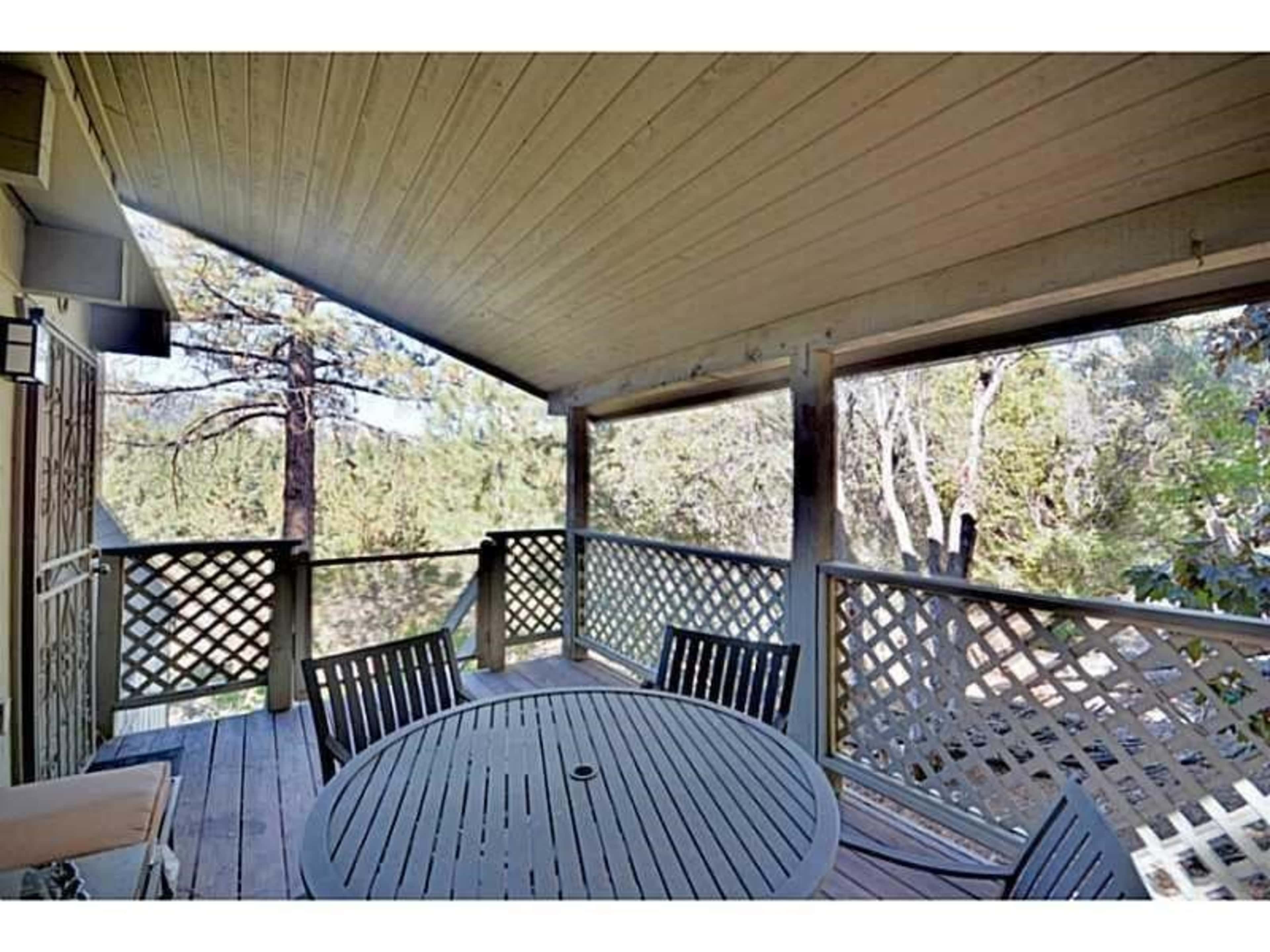 A covered wooden deck features a circular table with four chairs and overlooks a forested area.