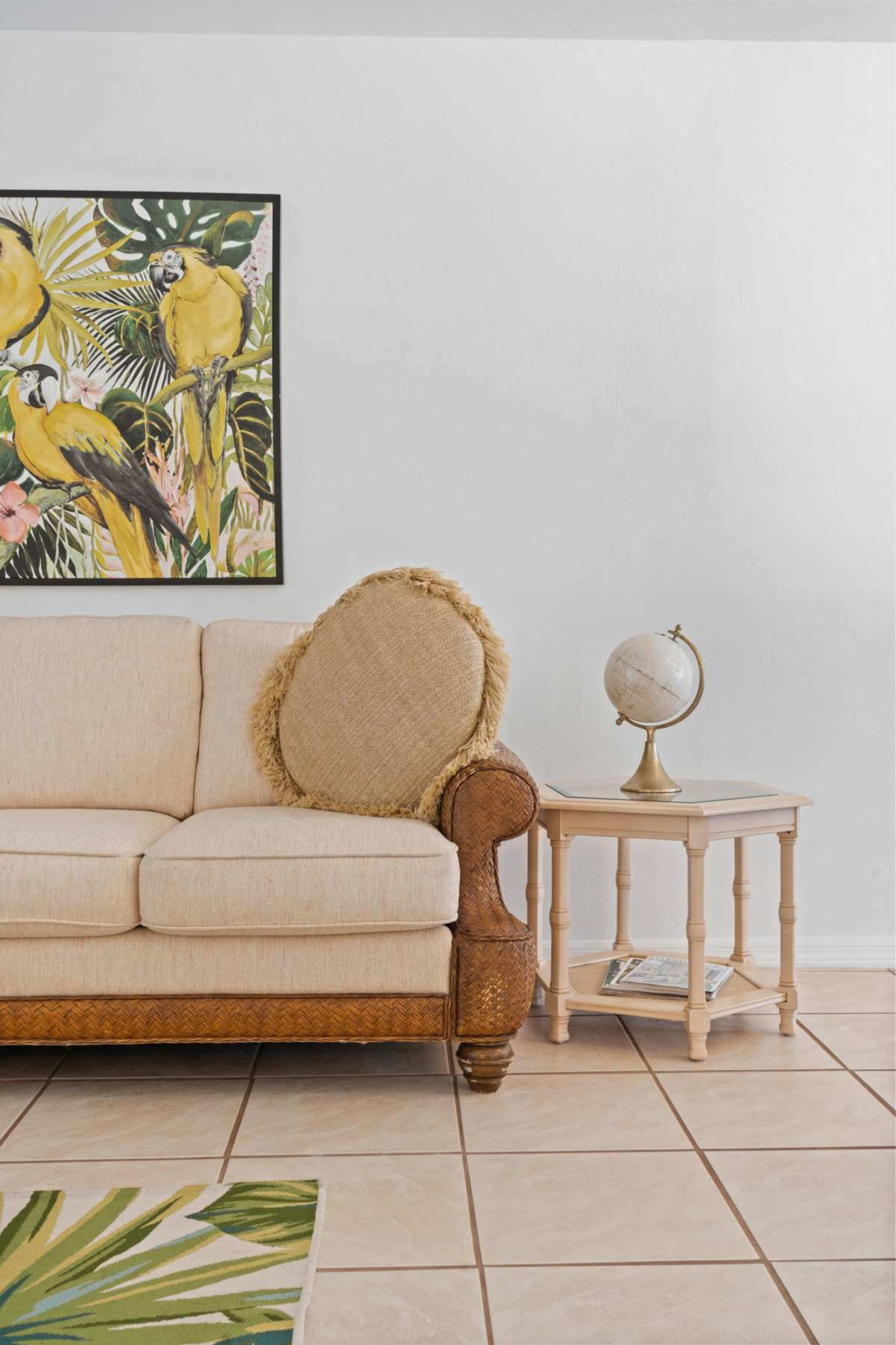 A beige sofa with a round, textured pillow sits next to a small table with a globe, all framed by a vibrant wall art featuring tropical birds.