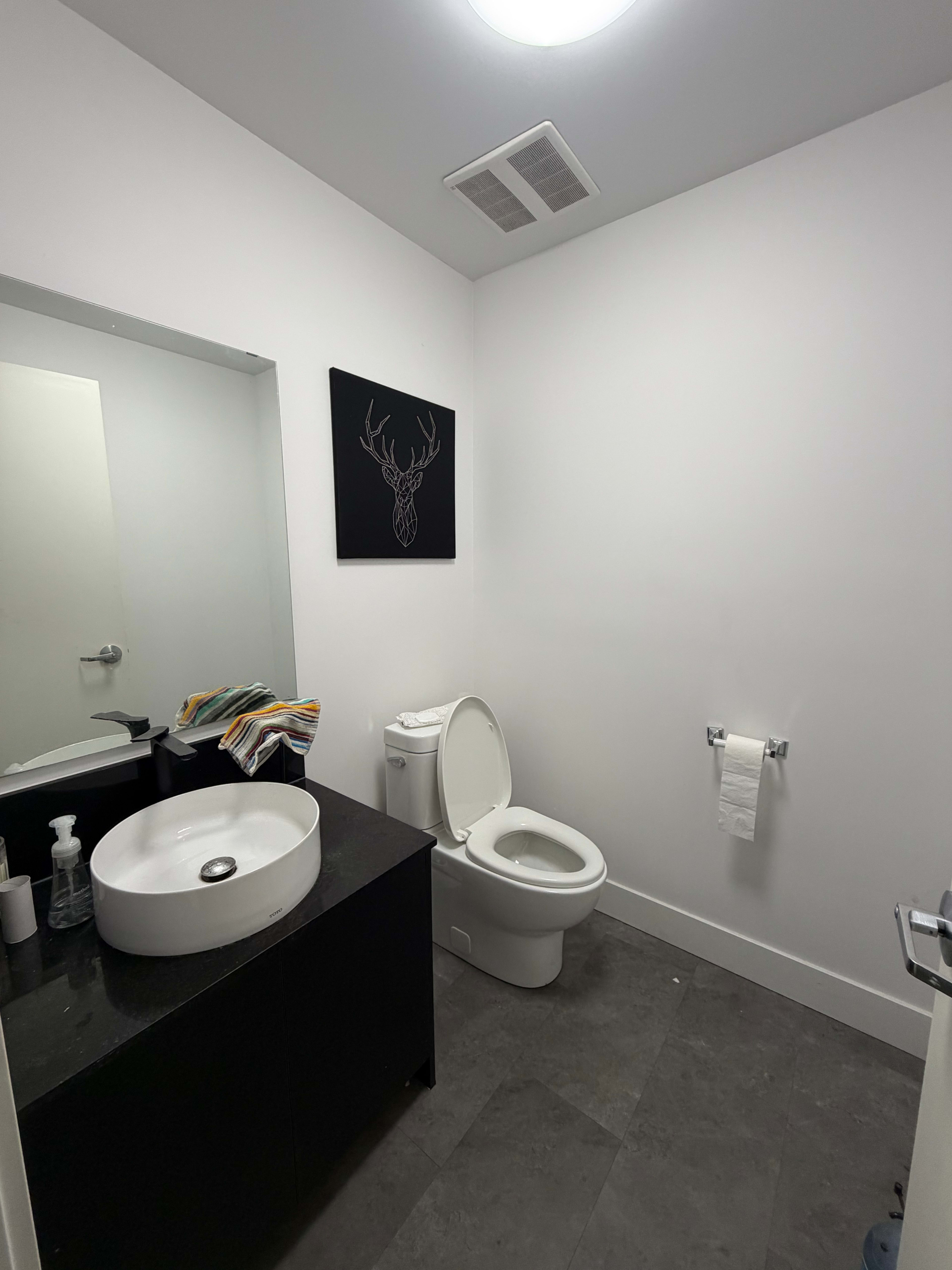 The image shows a modern bathroom featuring a toilet, a round sink on a dark vanity, a wall-mounted towel holder, and a minimalist deer head decoration.