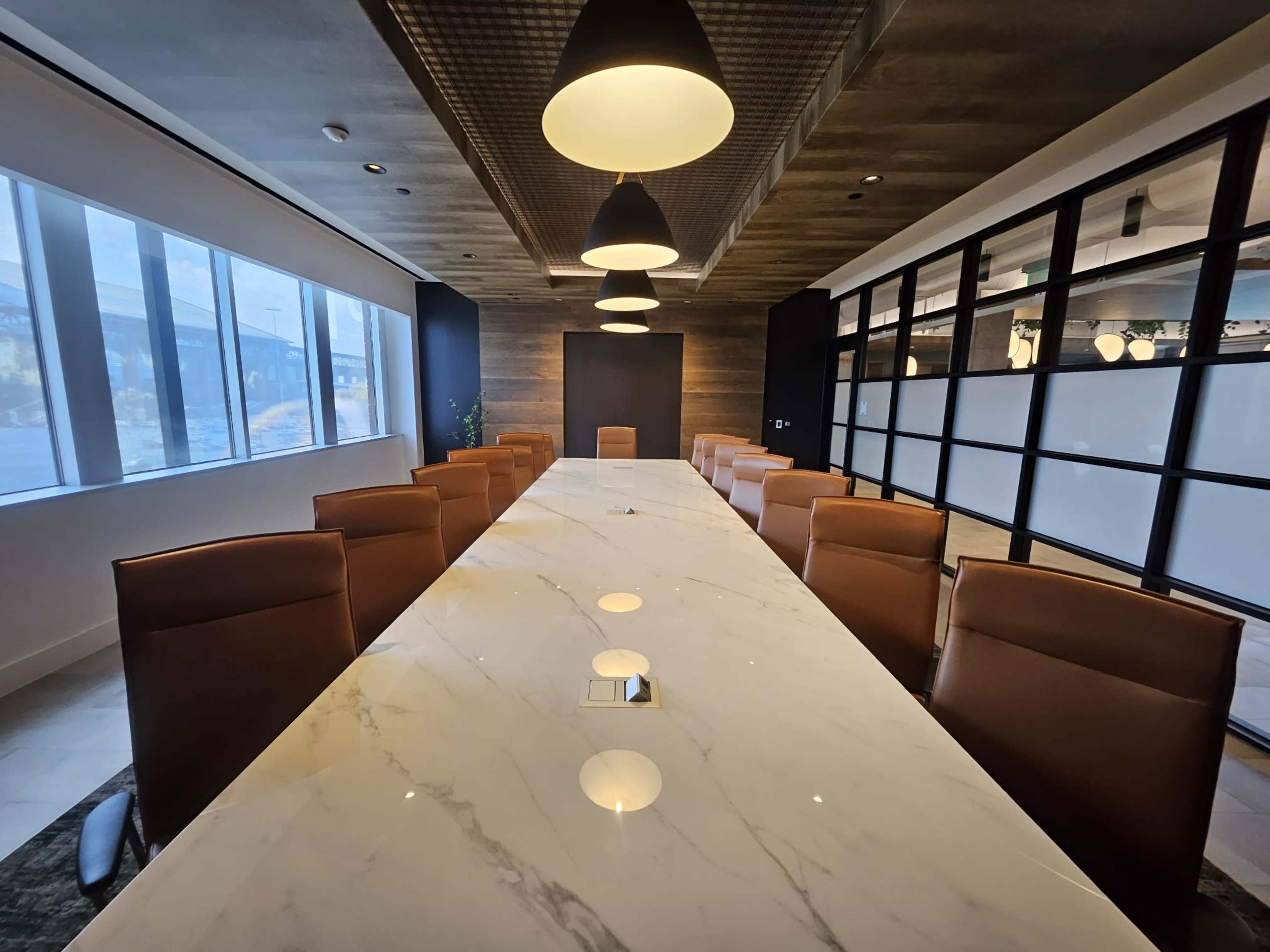 A long marble conference table with brown leather chairs is set in a modern meeting room illuminated by pendant lights.