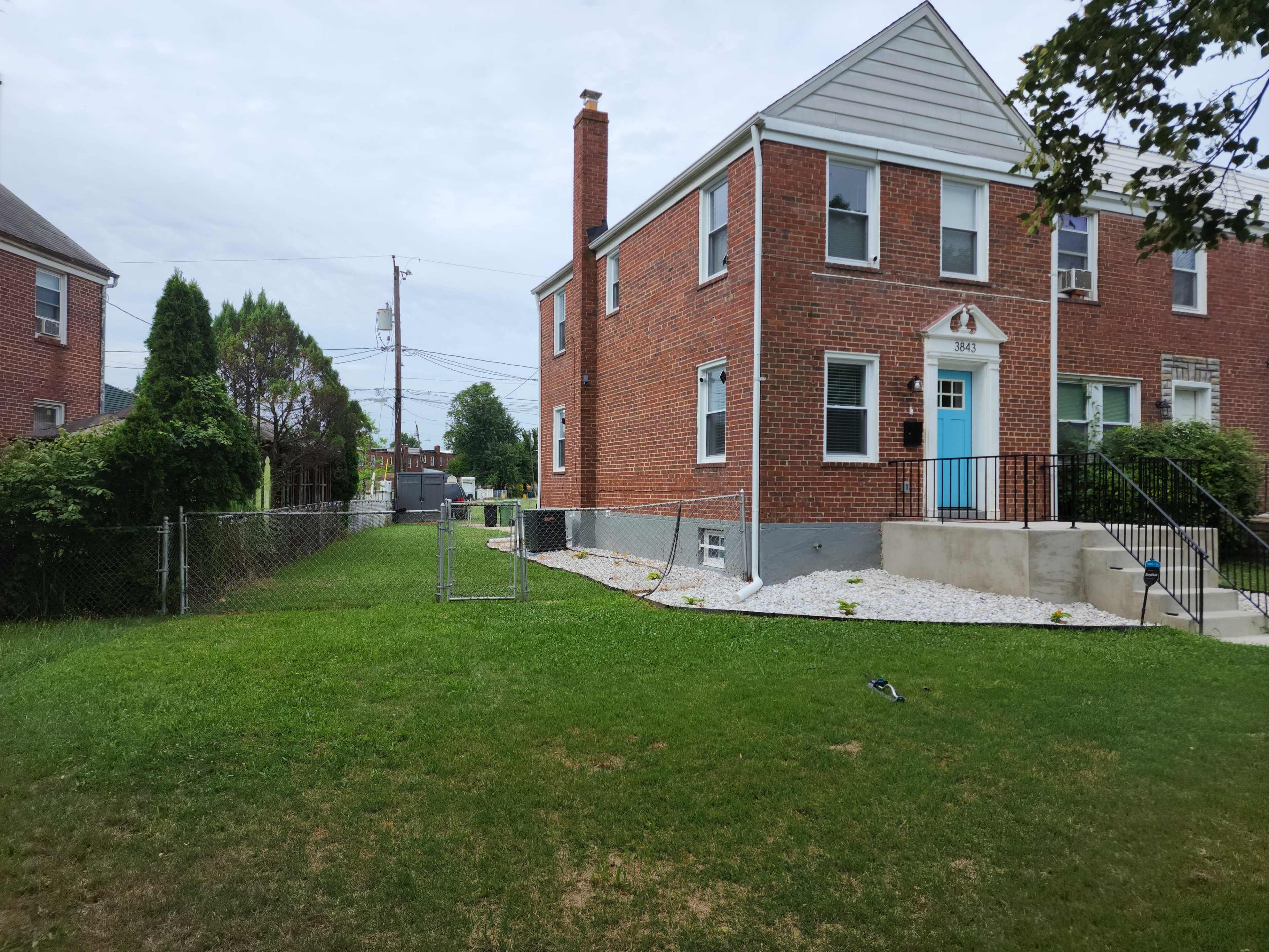 The image shows a two-story brick house with a light blue door, a fenced yard, and a large grassy area.