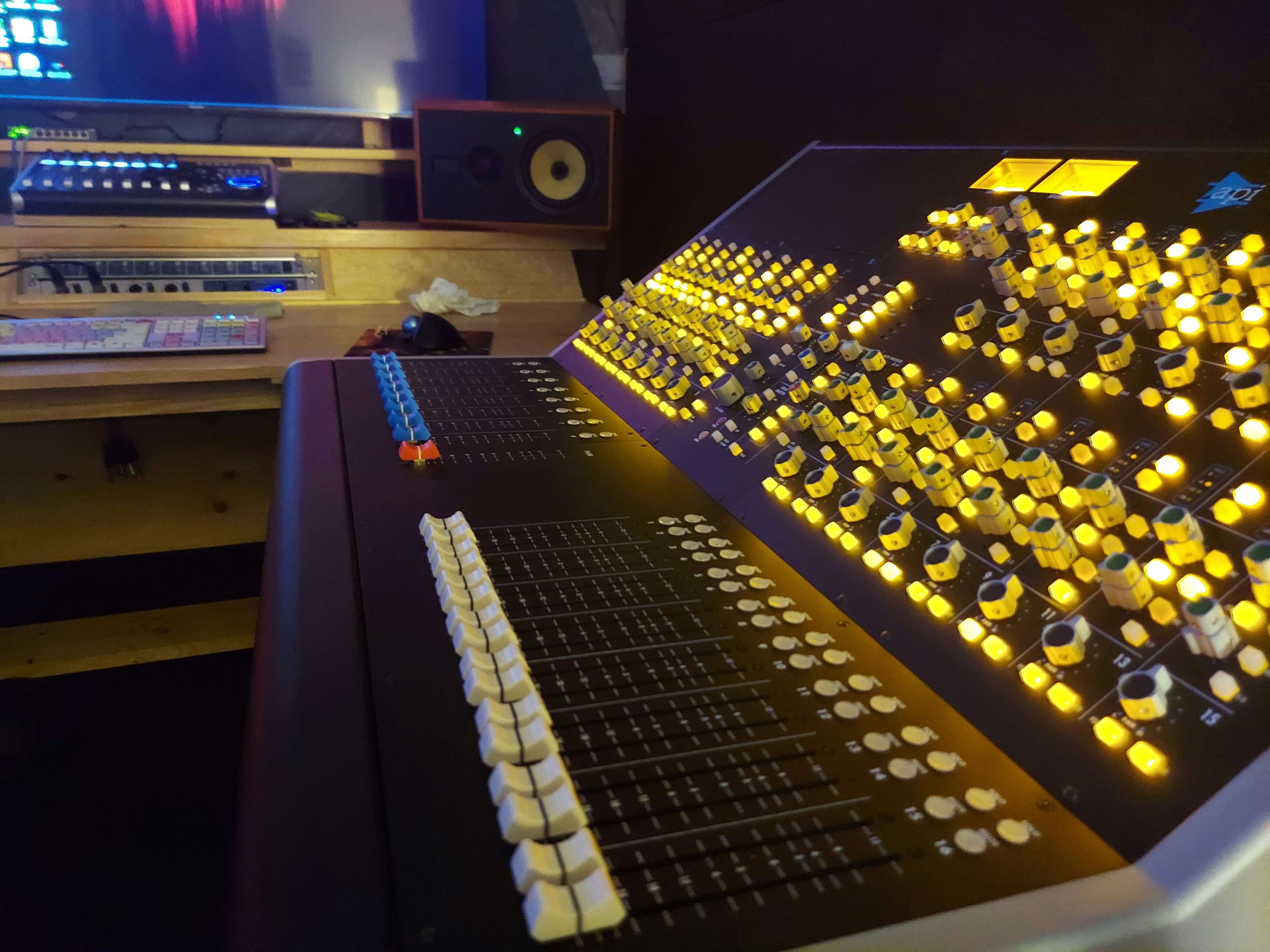 A close-up view of a recording studio's mixing console with illuminated buttons and faders, alongside a computer and monitors in the background.