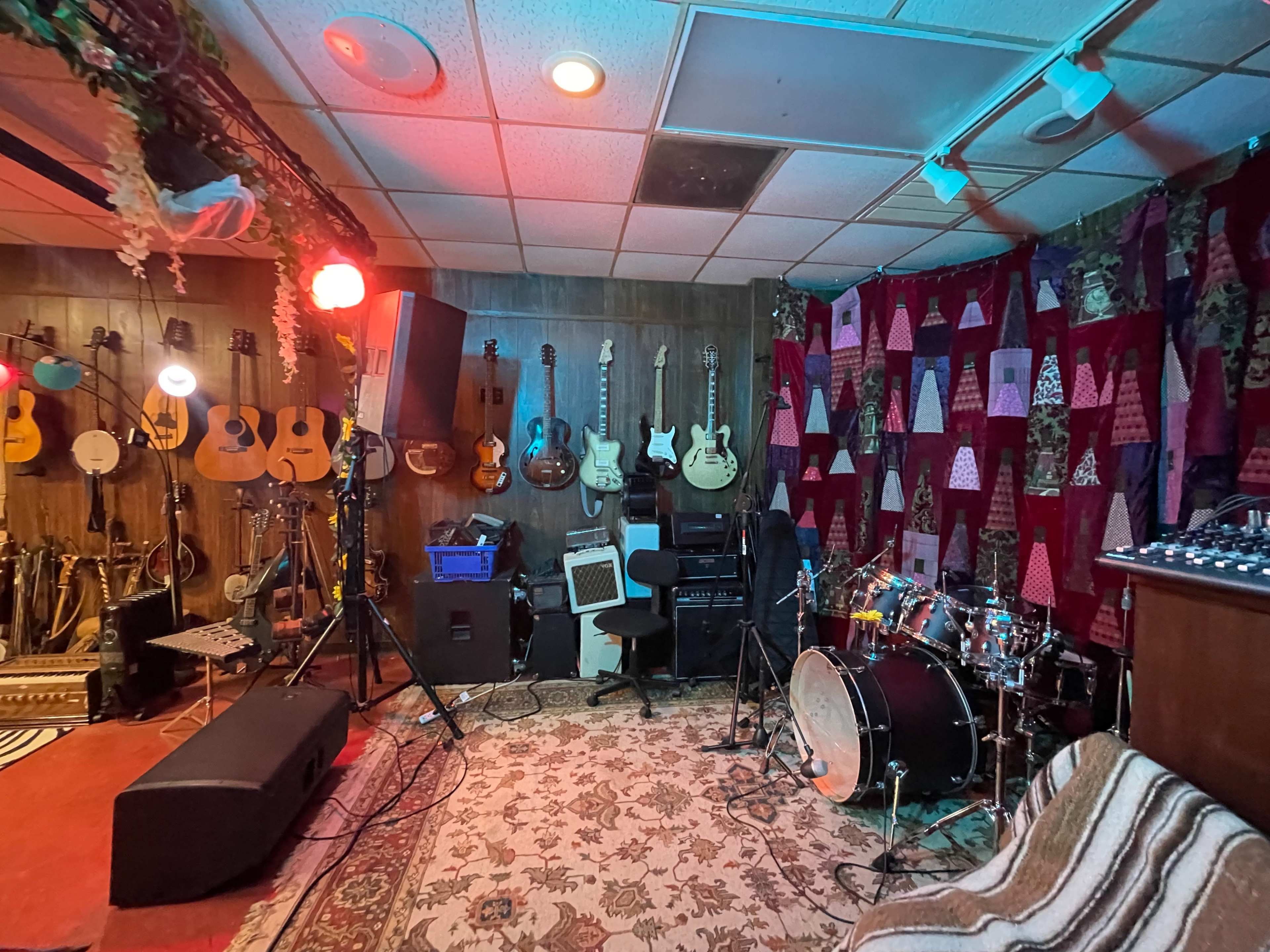 A music studio with various guitars hanging on the wall, a drum set on a patterned rug, and audio equipment set up for recording.