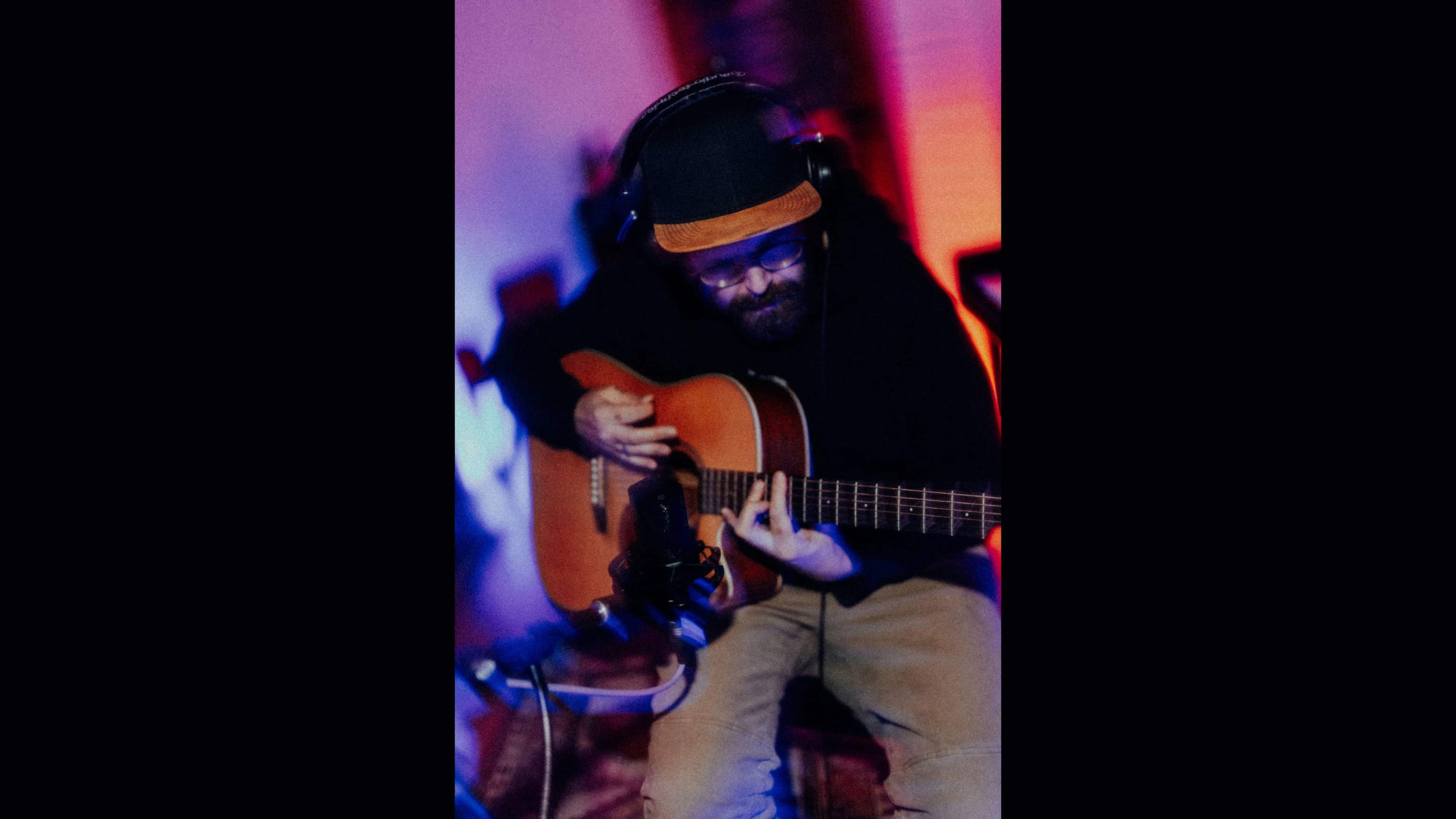 A musician plays an acoustic guitar while wearing headphones in a dimly lit room with colored lighting.