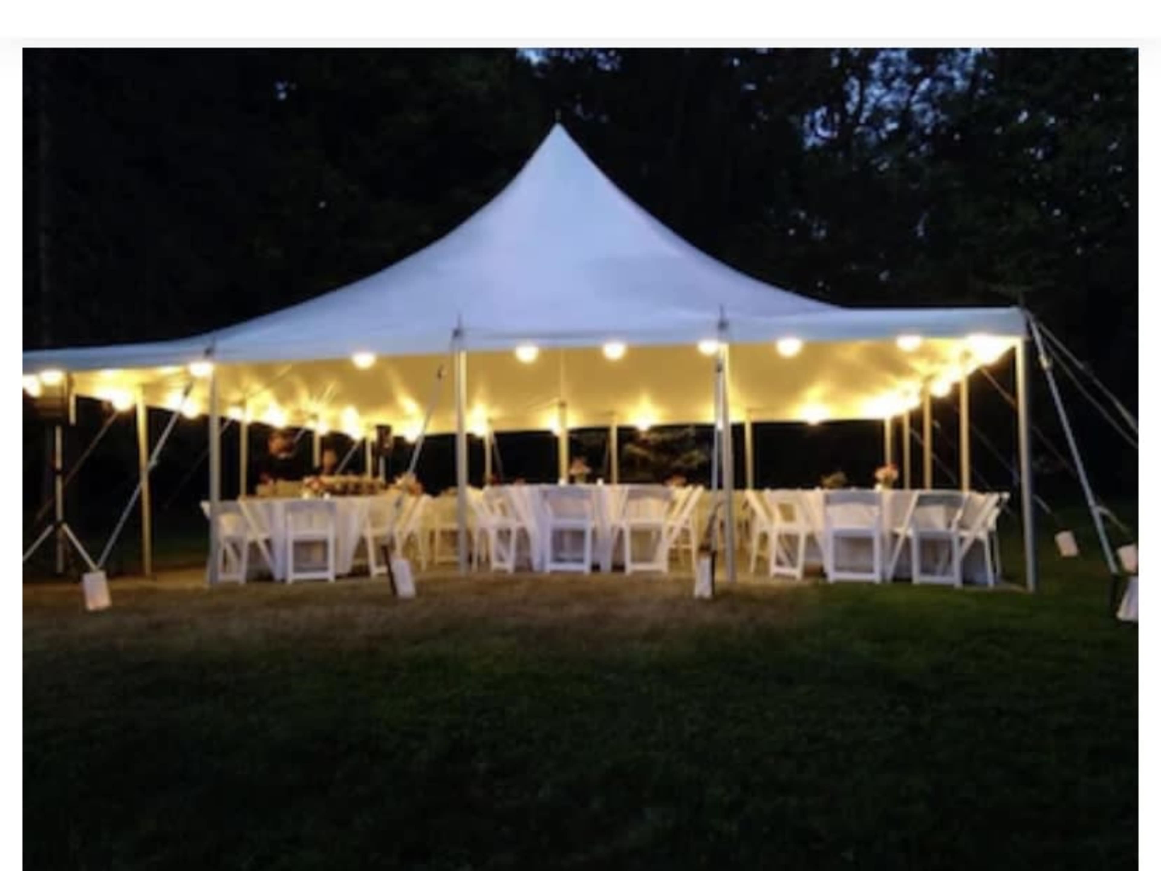 A large white tent with string lights is set up outdoors, featuring several tables and chairs arranged beneath it.