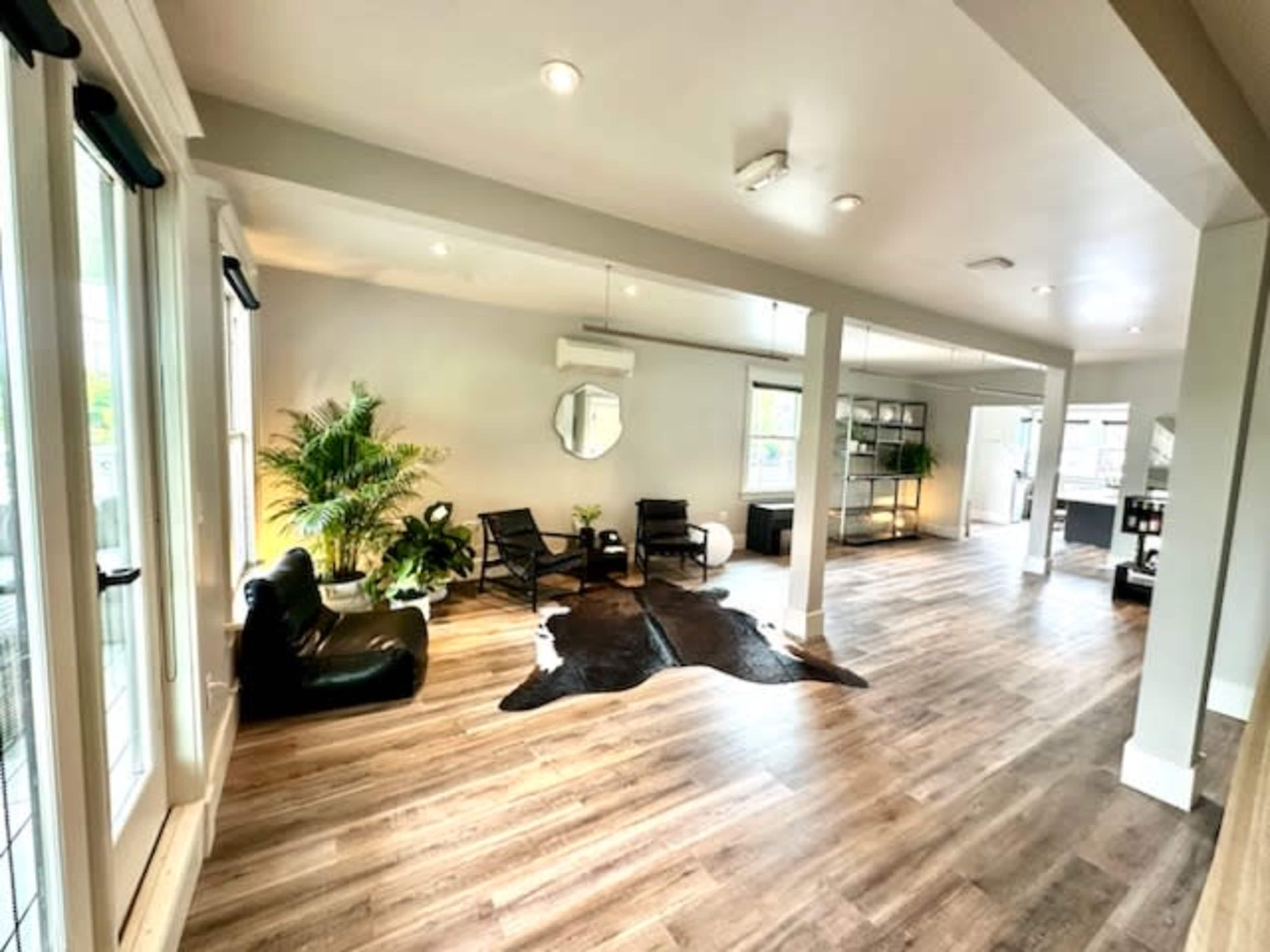 A spacious room with wooden flooring, featuring a seating area with black chairs, a large circular mirror, a plant, and a decorative cowhide rug.