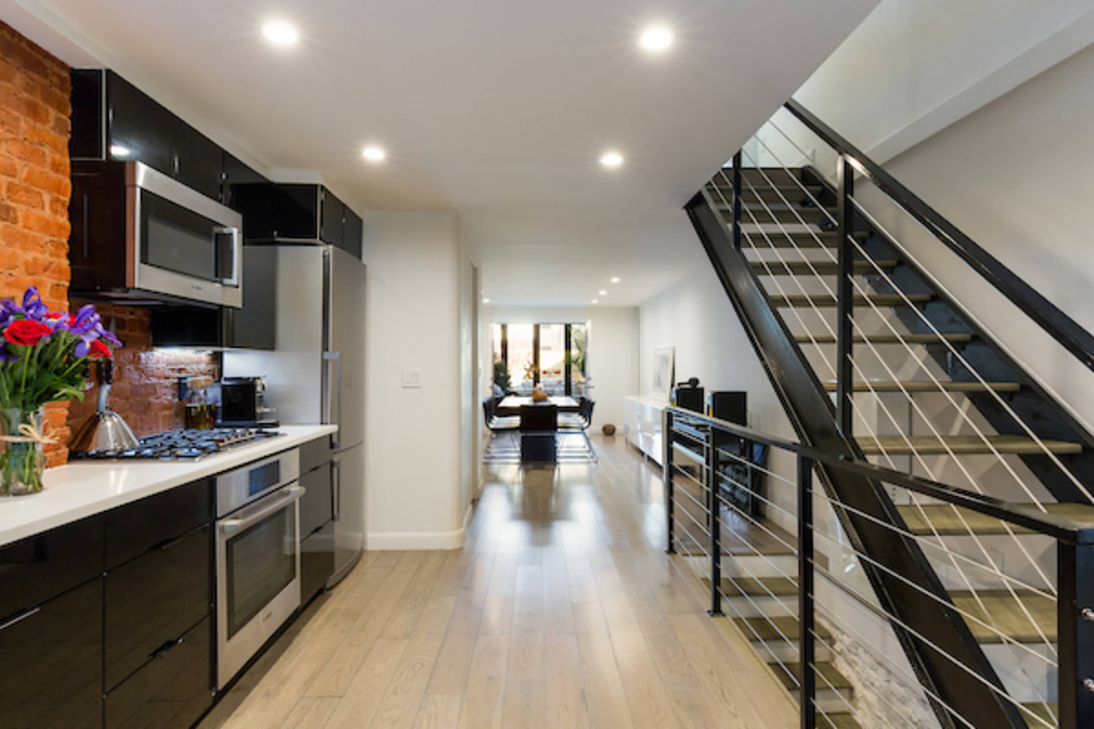 A modern kitchen with black cabinetry and stainless steel appliances, leading to a hallway with a staircase and large windows at the far end.