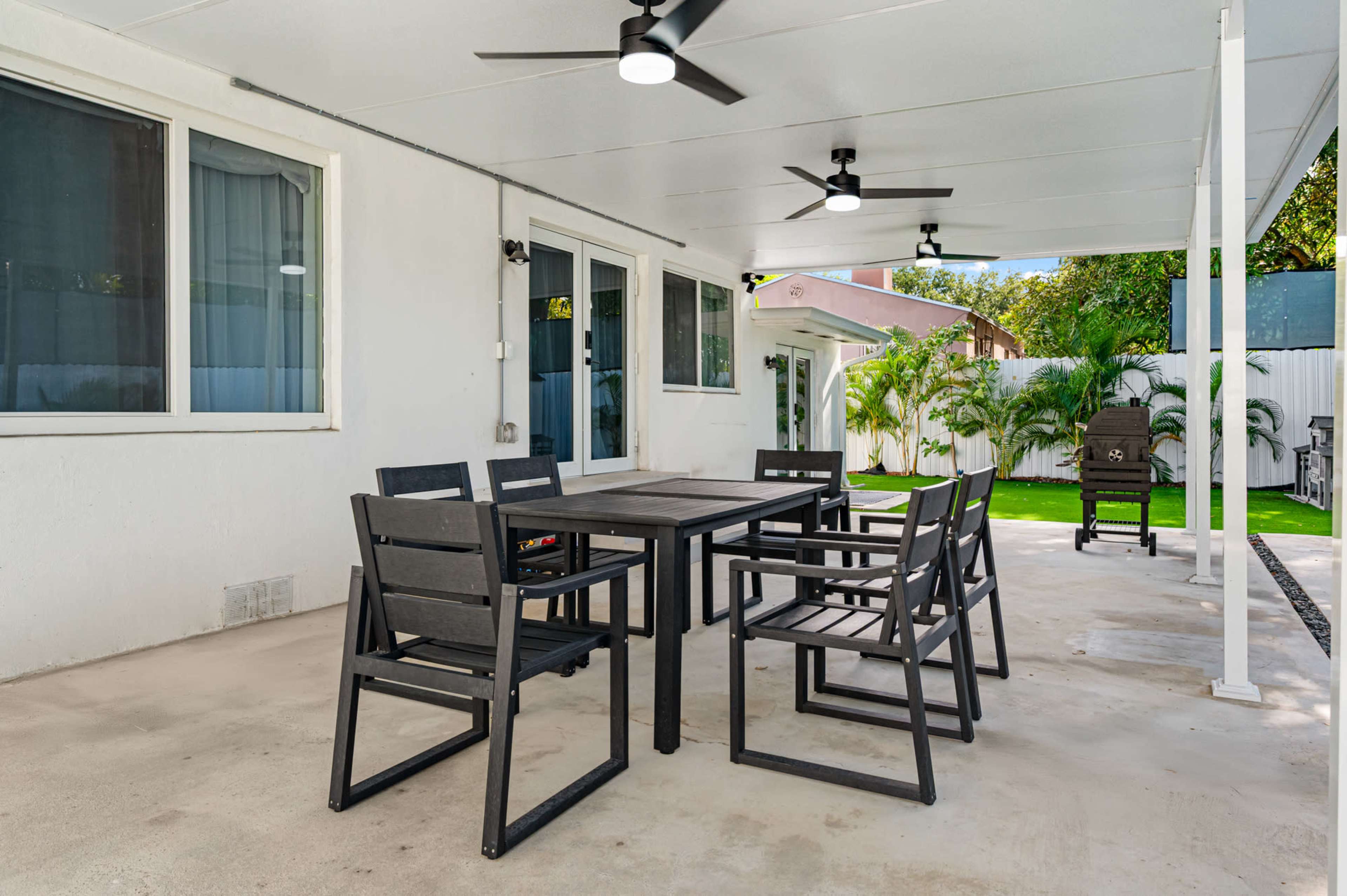 The image shows a covered patio with a wooden dining table and six chairs, surrounded by tropical plants.