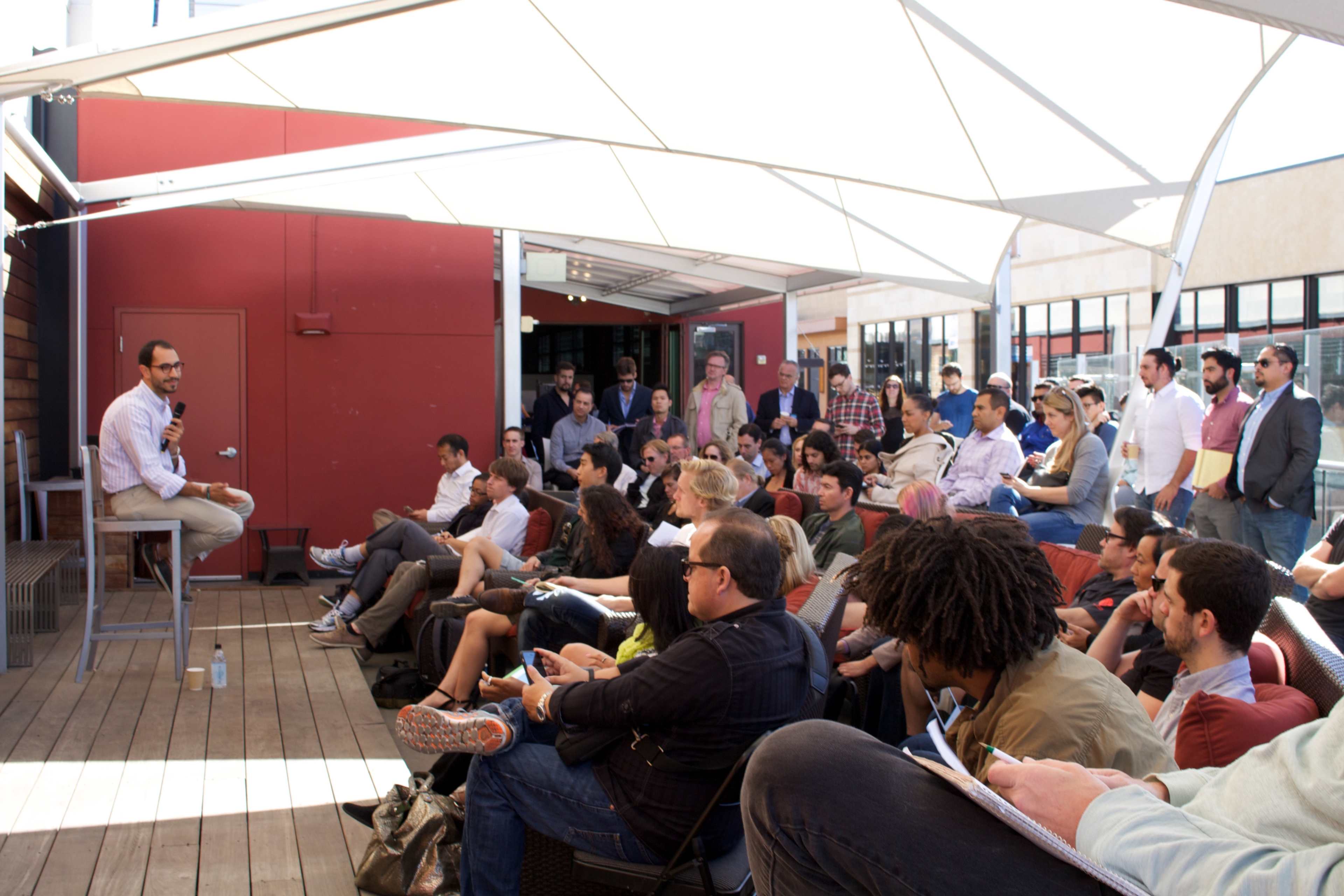A speaker sits on a stool addressing a large crowd in a rooftop event space.