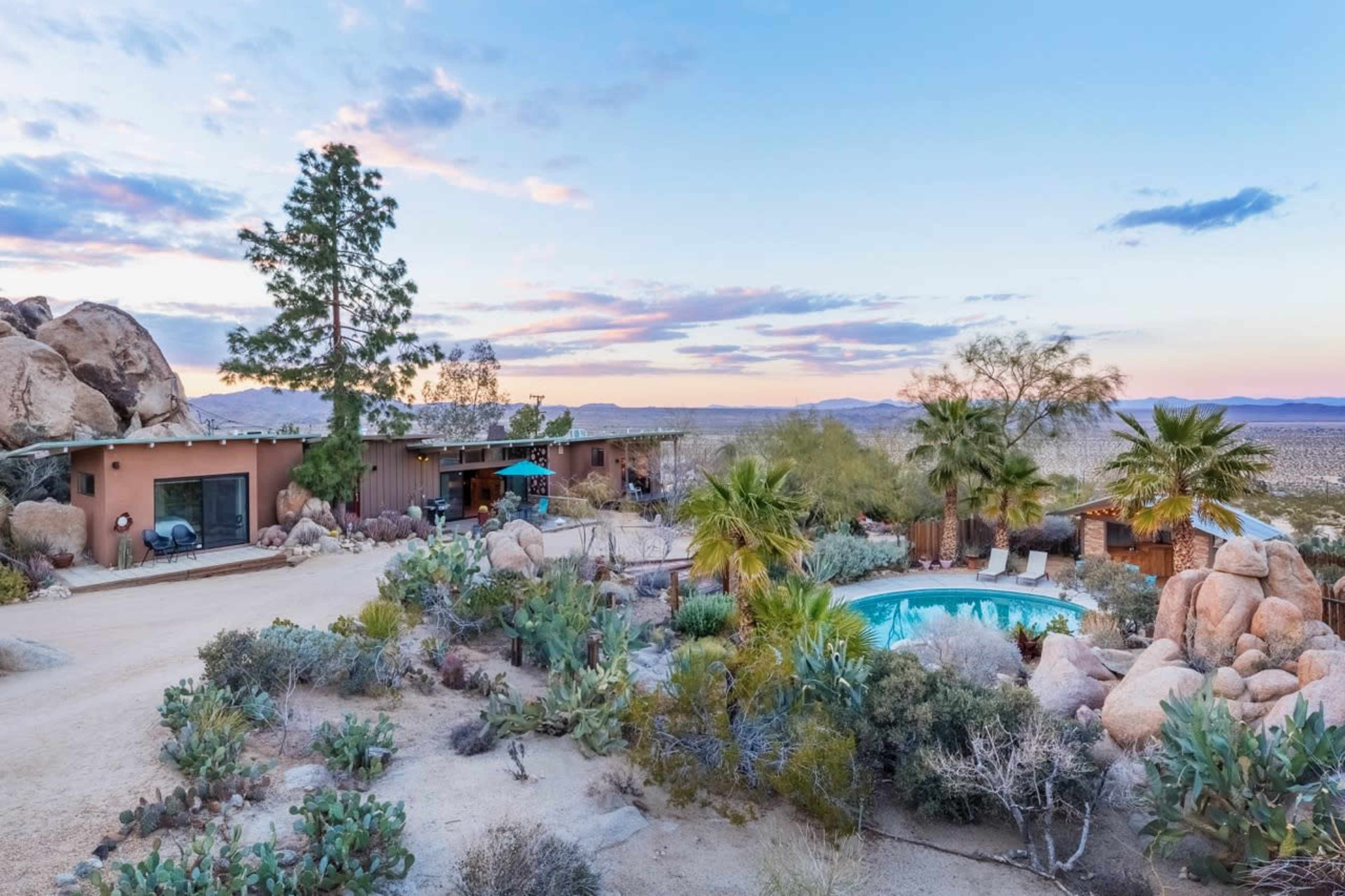 A desert landscape featuring a modern house with a swimming pool surrounded by large rocks and desert vegetation.