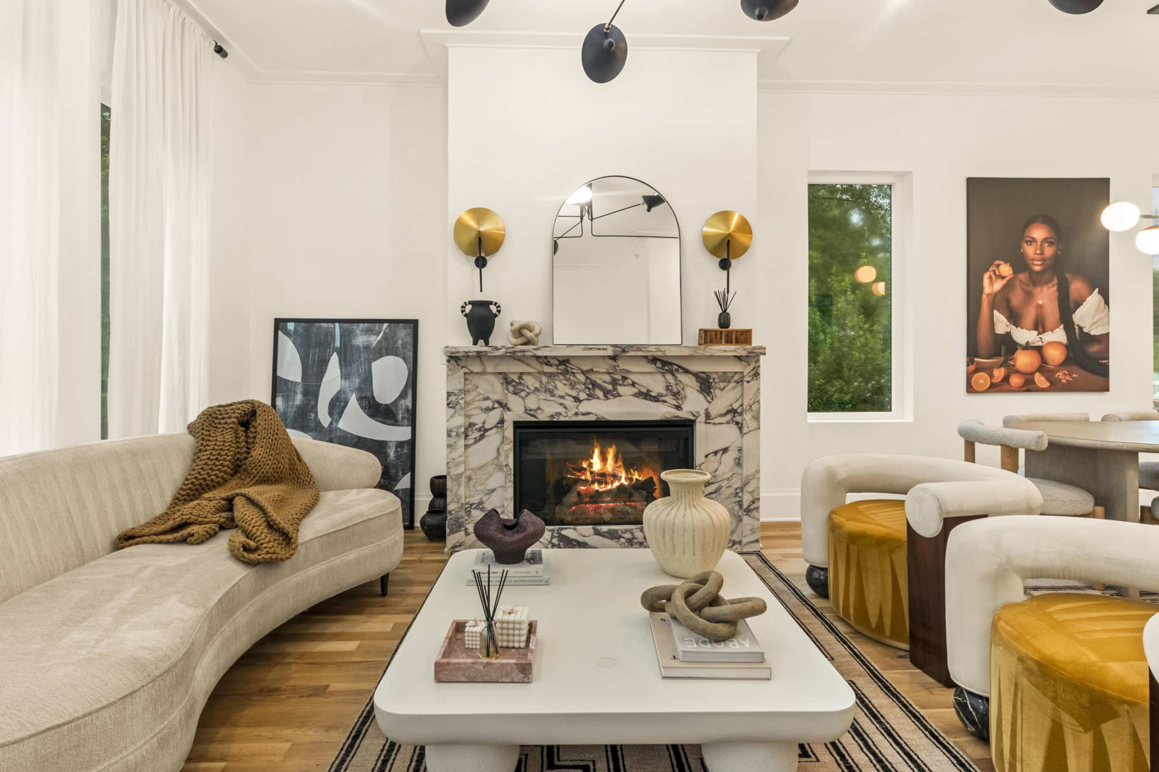 The image shows a modern living room featuring a curved sofa, a marble fireplace with a mirror above it, and a dining area with a table and yellow upholstered stools.