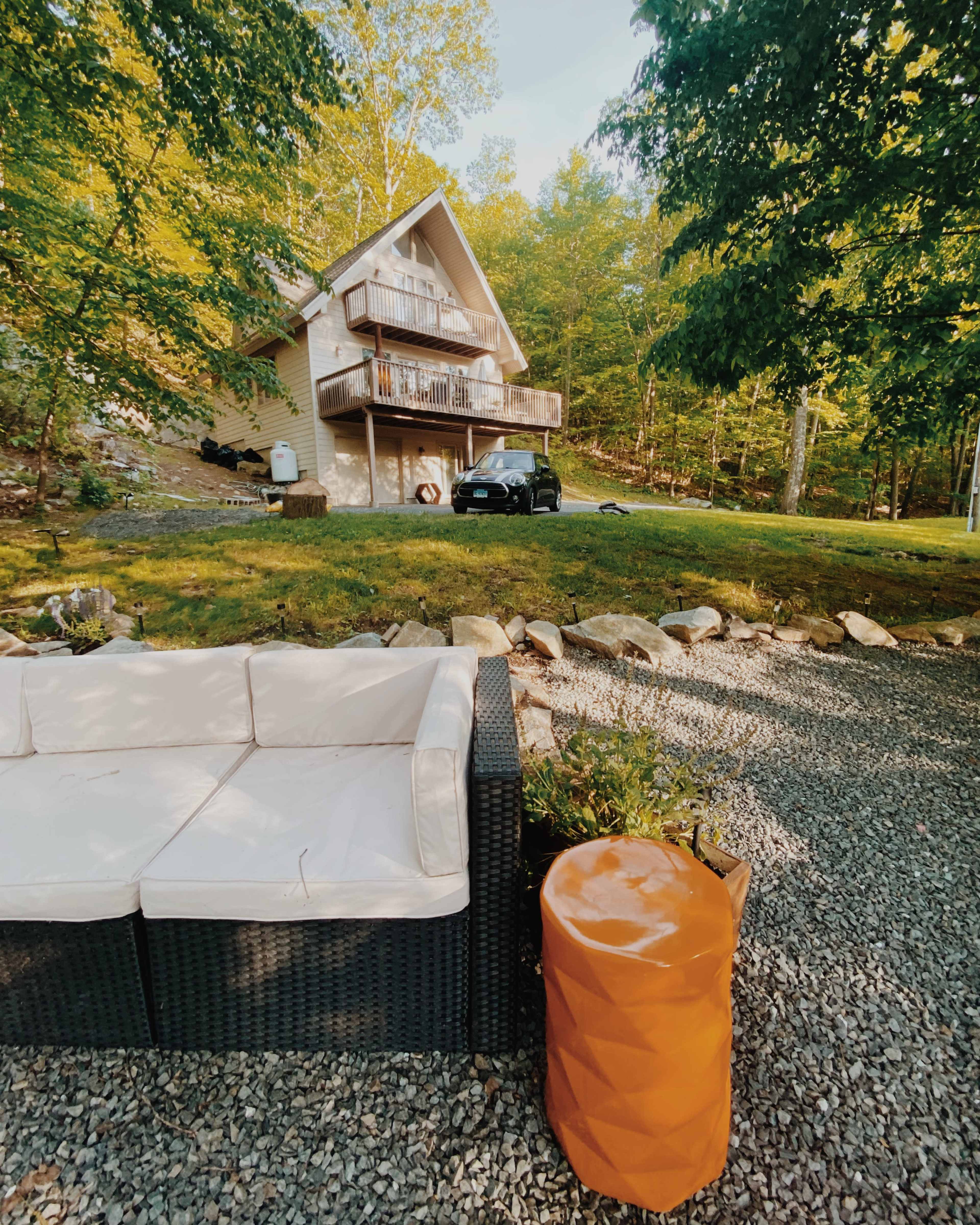 A two-story wooden house is situated on a grassy area surrounded by trees, with a modern couch and an orange side table in the foreground on a gravel surface.