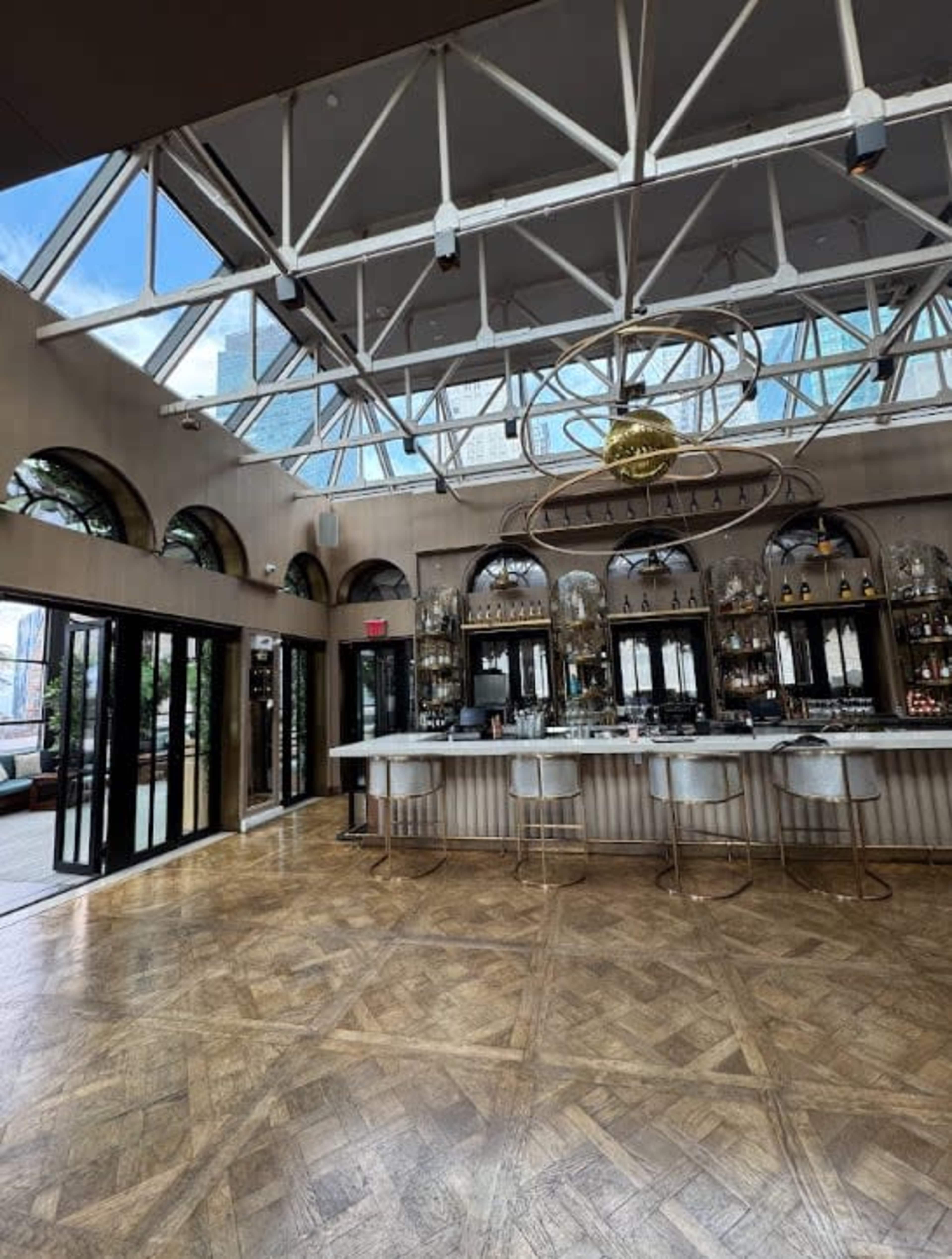 The image shows a modern bar area with a high glass ceiling, a polished wooden floor, and a well-stocked bar featuring circular stools.