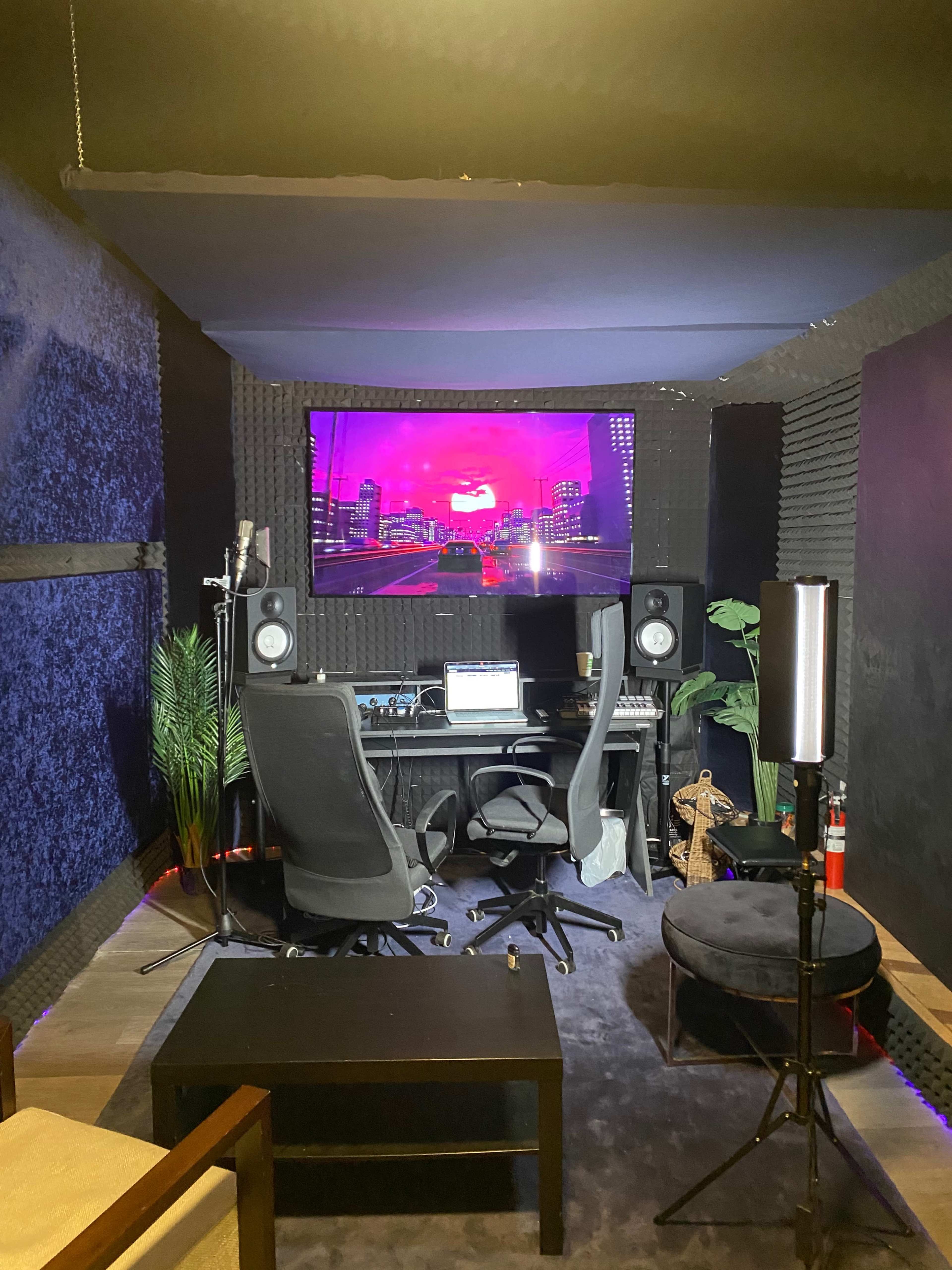 A home recording studio featuring two chairs, a desk with audio equipment, a large screen displaying a colorful cityscape, and soundproofing panels on the walls.