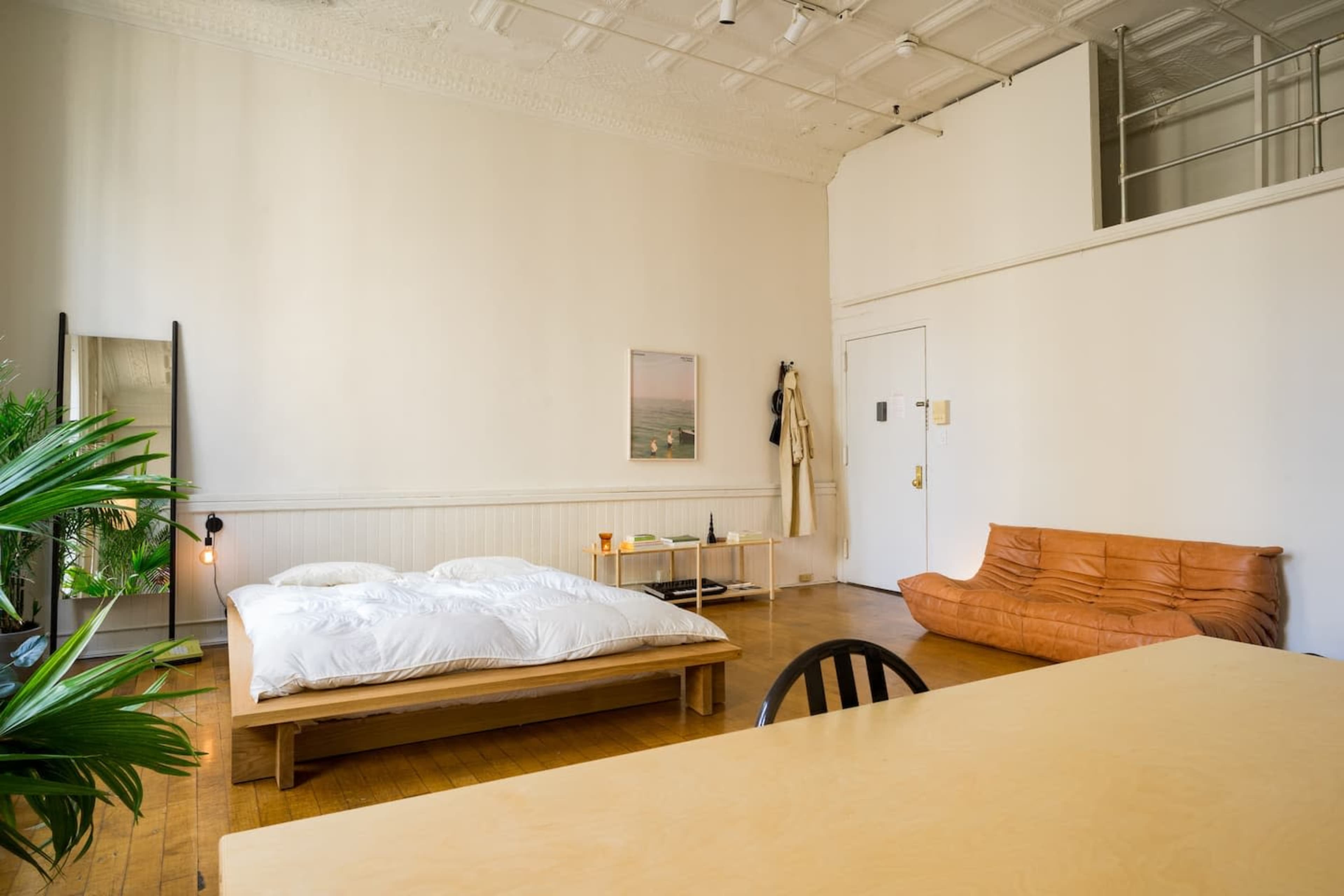 The room features a bed with white bedding, a wooden table, a brown sofa, a large mirror, and plants in a minimalistic layout.
