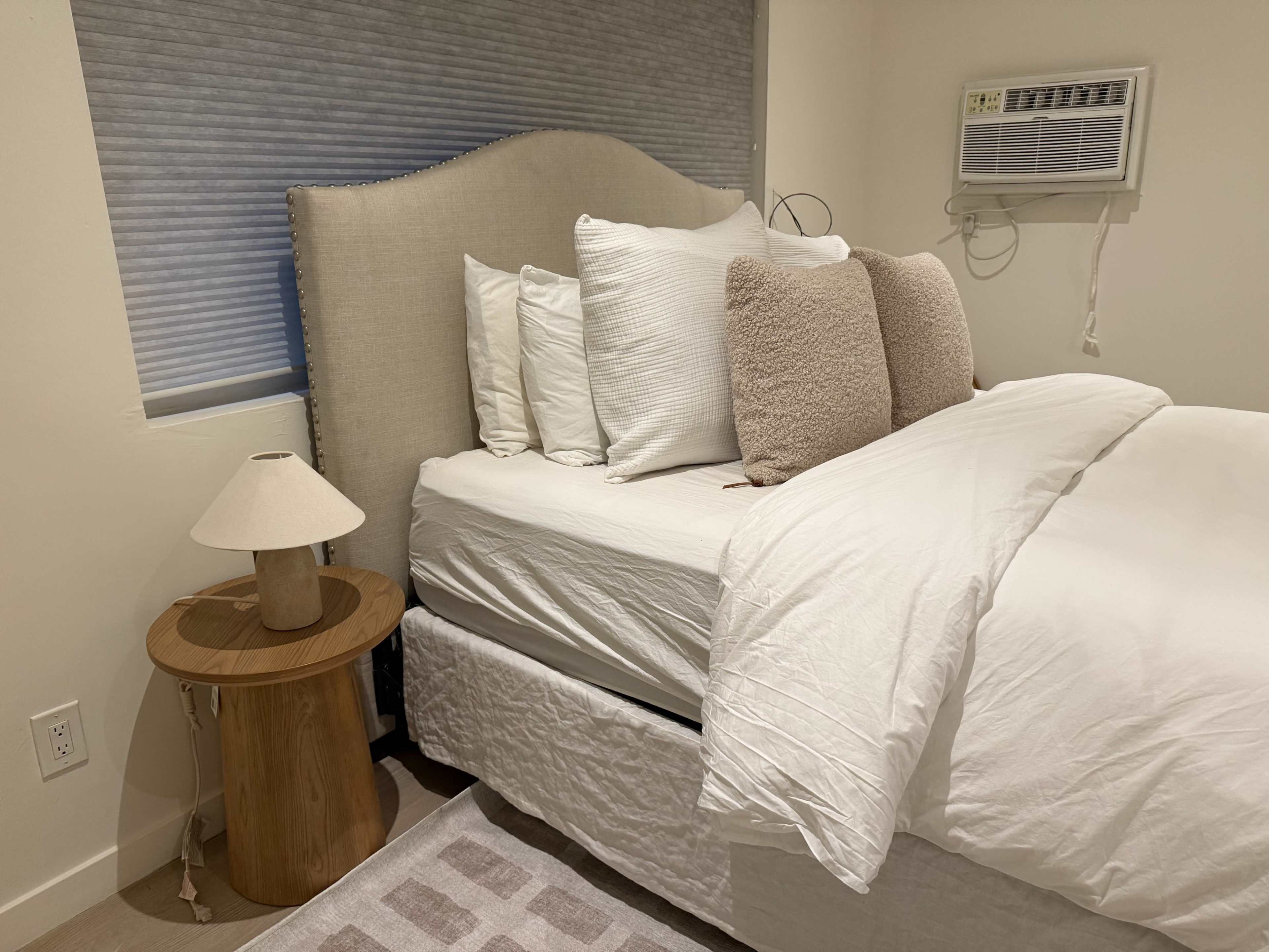 A neatly made bed with white bedding and multiple pillows is situated next to a small wooden side table and a lamp in a softly lit bedroom.